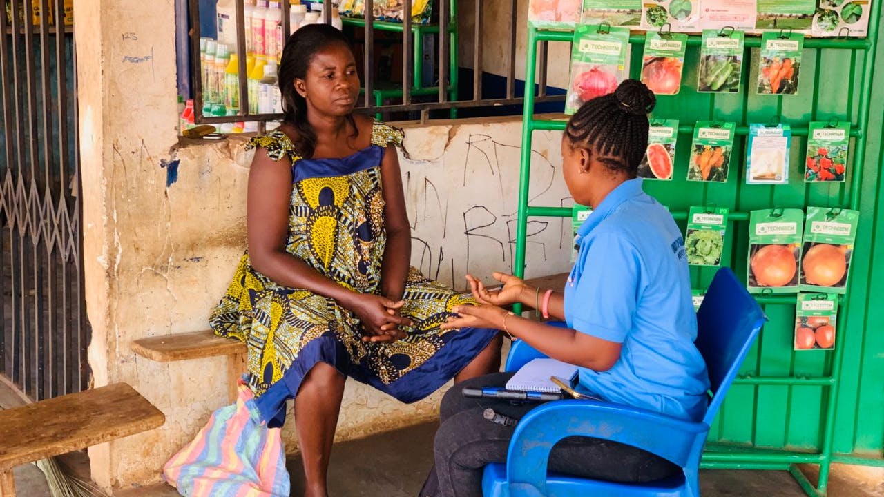 Kalian Sampoa Gumah conducts a survey with a female farmer. The farmer wears a brightly coloured dress. The are seated outside a small grocery store next to a green metal stand with rows of seeds in packets. (Ghana, 2025)