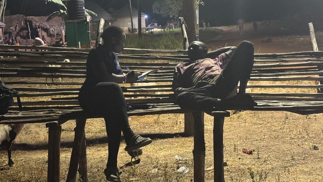 It is nighttime. Kalian Sampoa Gumah sits outside on a makeshift bench and conducts a survey with a male farmer who is leaning back and relaxing, with his hand behind his head.