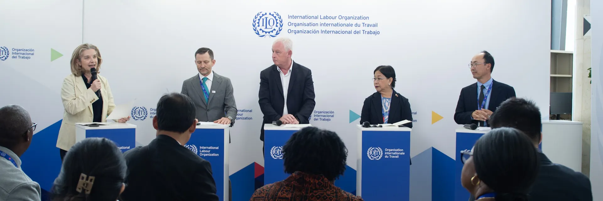 Five people stand behind lecterns, from left to right: a woman who holds a microphone, two men, another woman and lastly a man. In the foreground are people who are seated with their backs to the camera, facing the people at the lecterns.