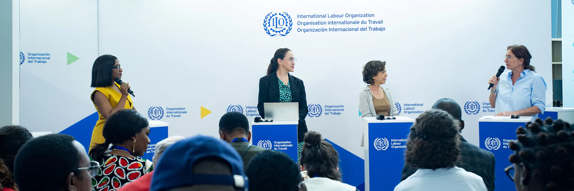 Four women stand behind lecterns and two hold a microphone. In the foreground are the backs of a seated crowd.