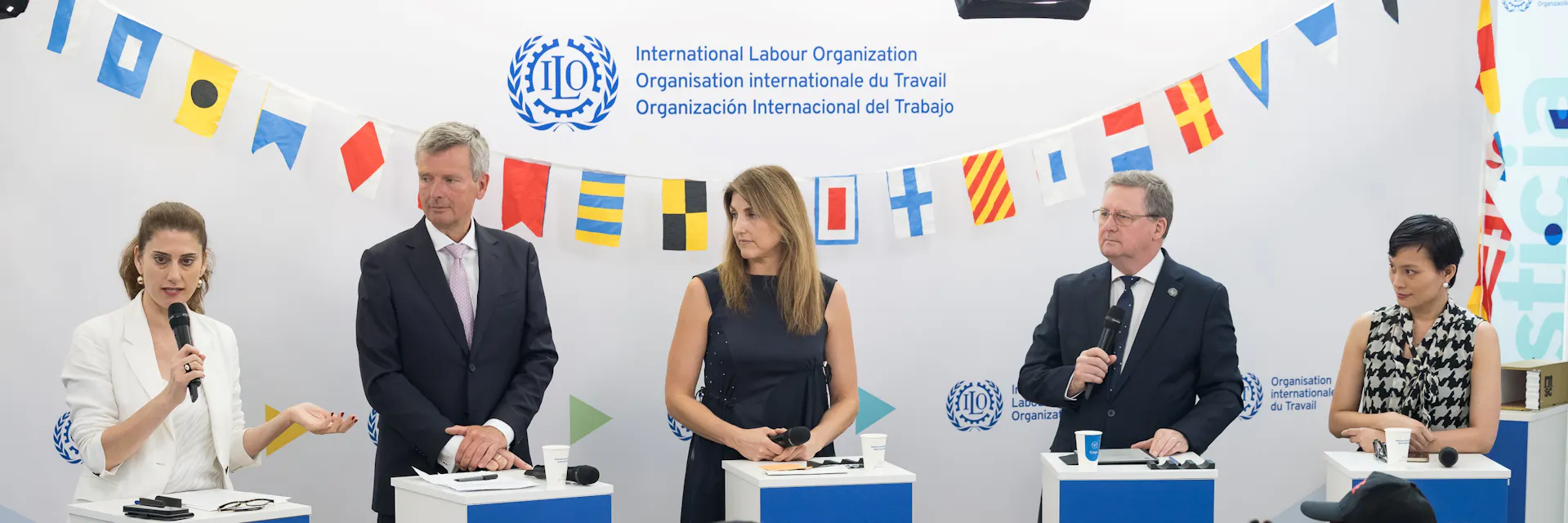 Five people - three woman and two men -stand behind lecterns. In the foreground a seated audience faces them. A string of flags from maritime states is on the wall behind the speakers.