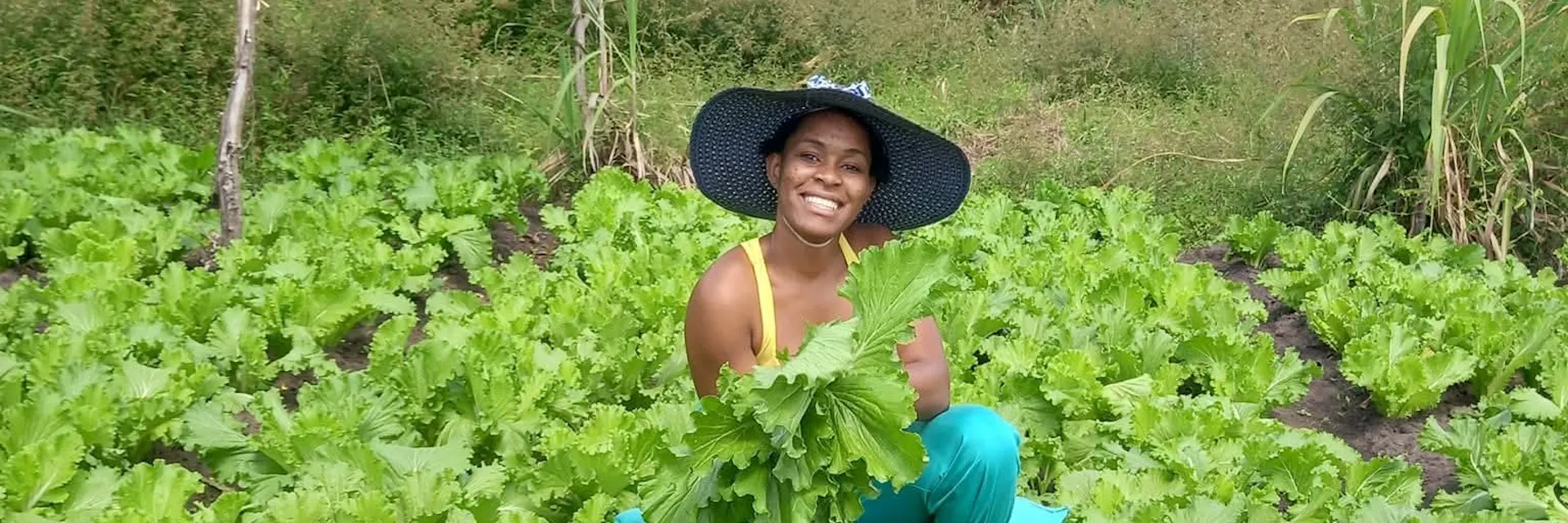Farmer Clara Kamlomo is crouched in a field of lettuce and holds a lettuce plant in her hands. She wears a hat and smiles at the camera.