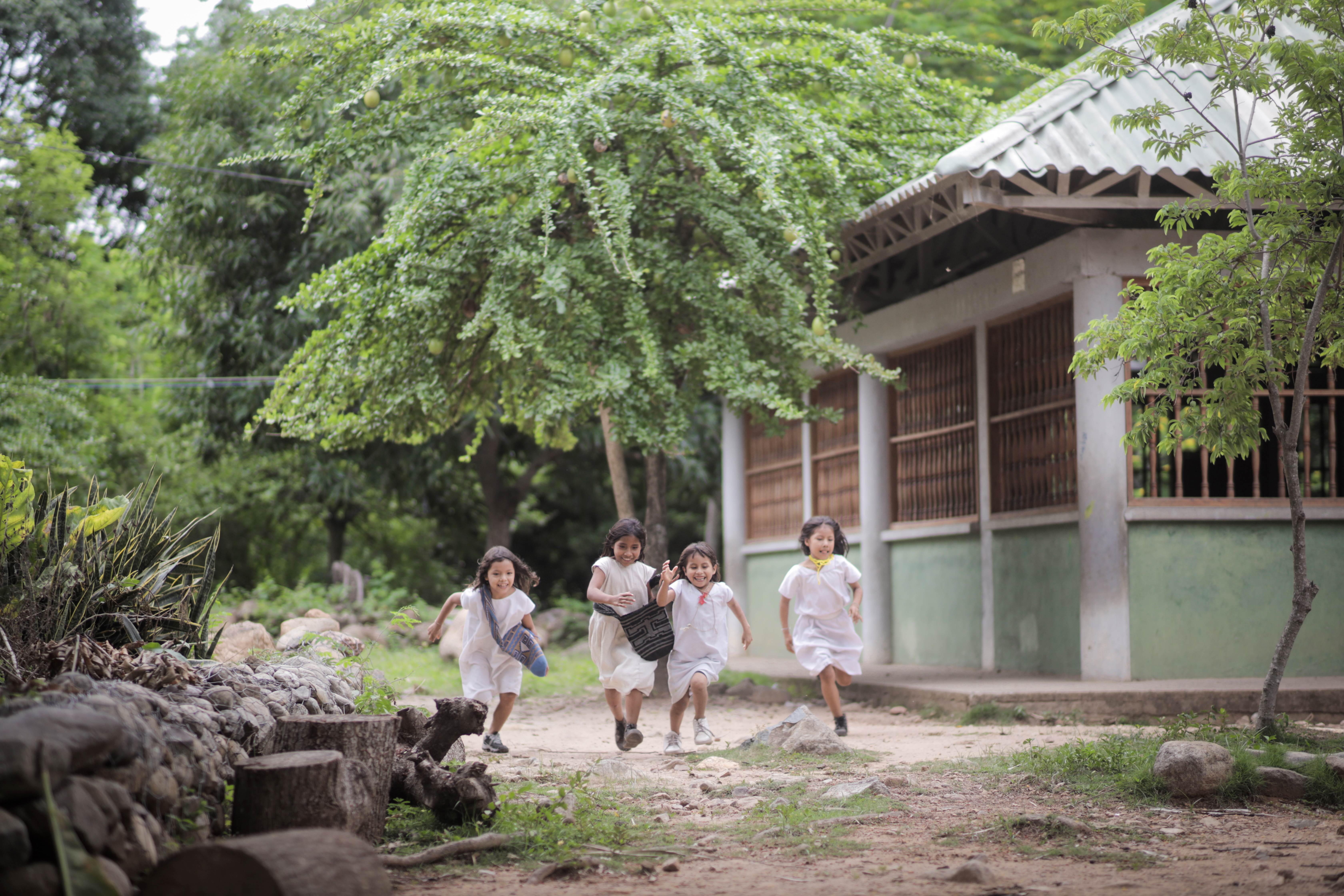 Children run towards the camera, laughing and carrying mochilla. In the background, there is a green building and trees.