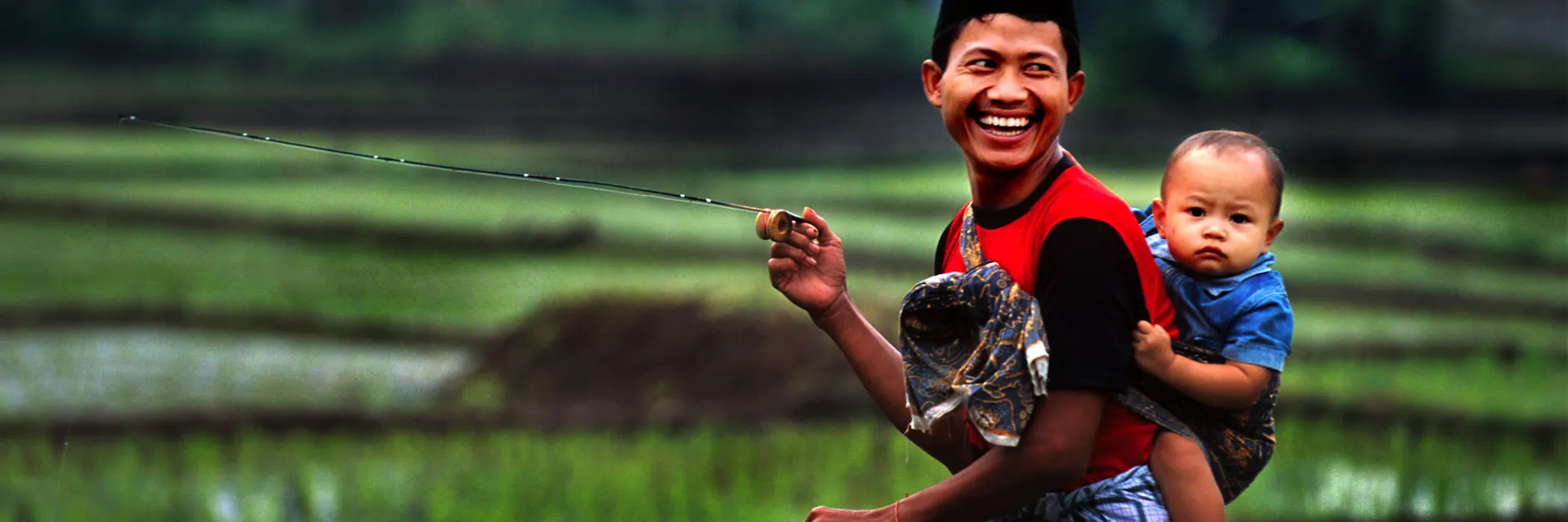 Un père porte son enfant sur le dos pendant qu'il pêche. Jakarta. 2015.