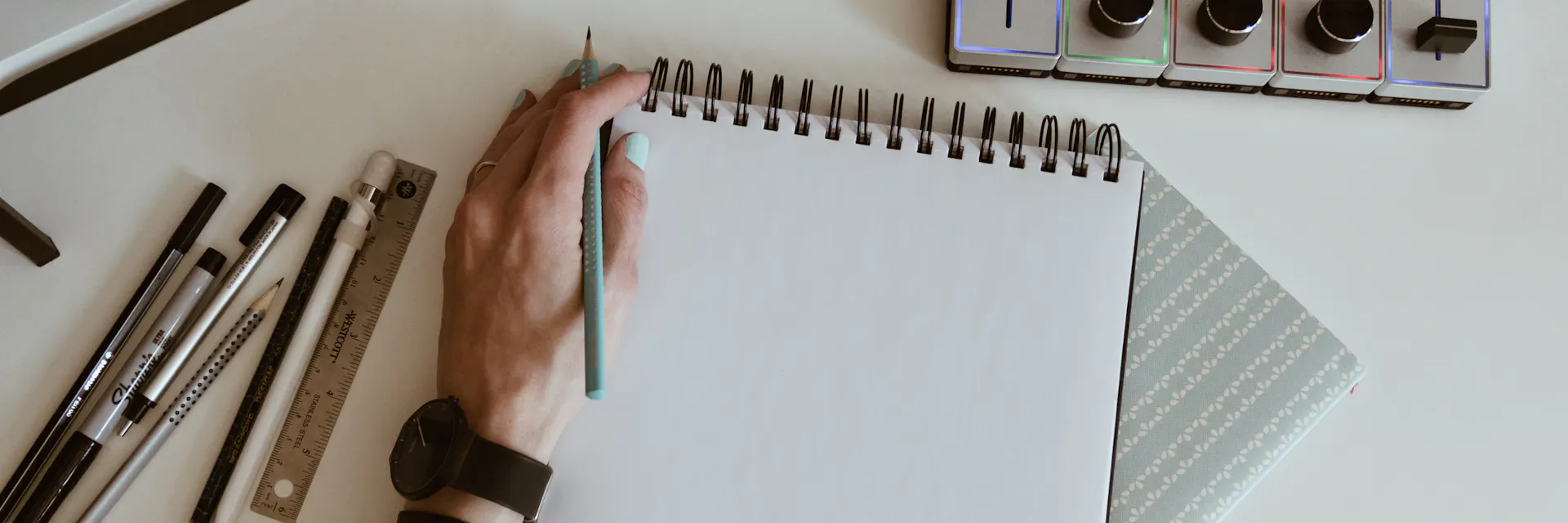 A left hand holds a pencil while resting on a notepad, while on the table around it lie more pencils and drawing tools.