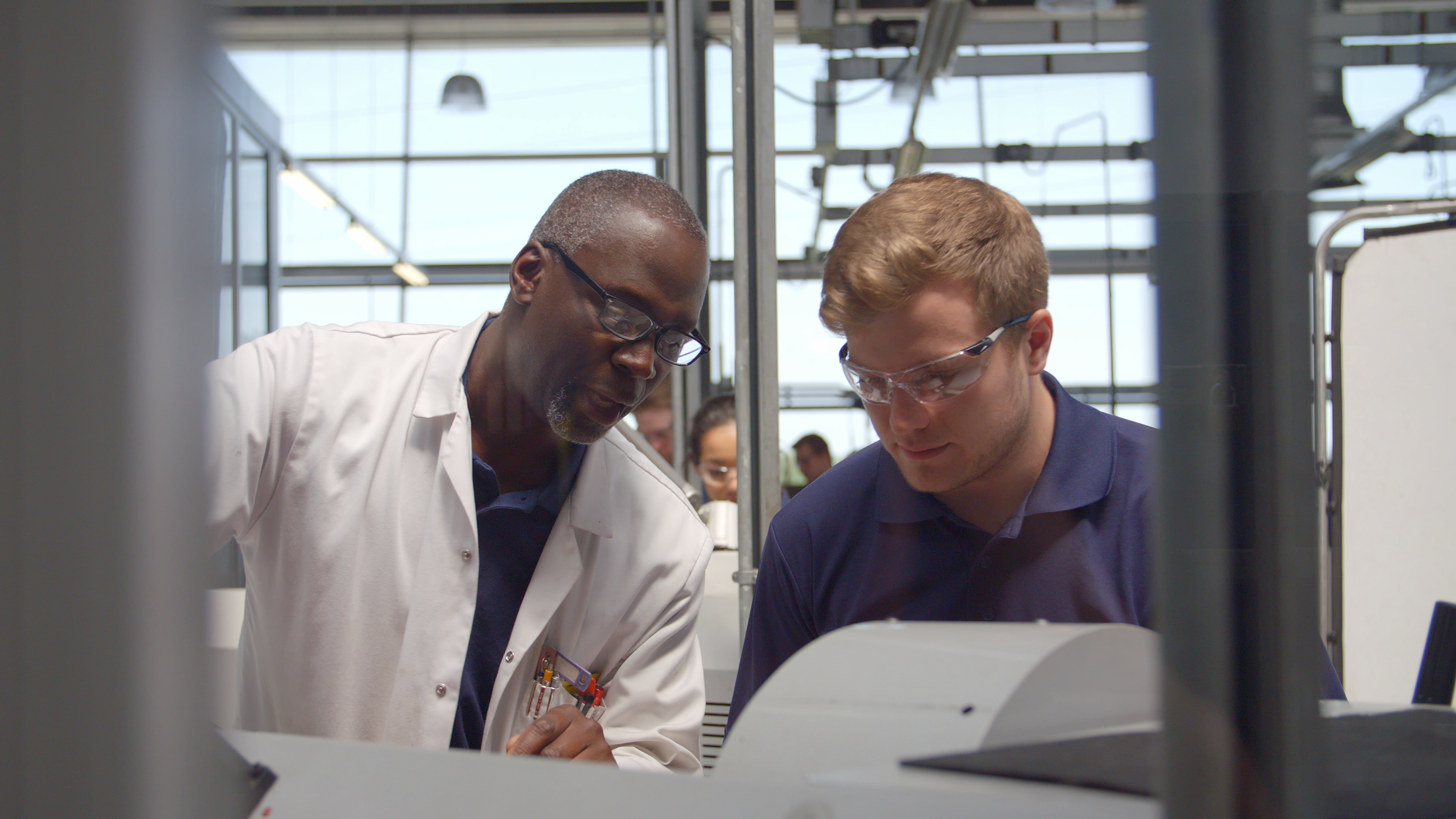 Two men who are engineers, one older on the left who wears a white lab coat, and one younger on the right, who is an apprentice and wears safety glasses, look down at something outside the frame of the image. 