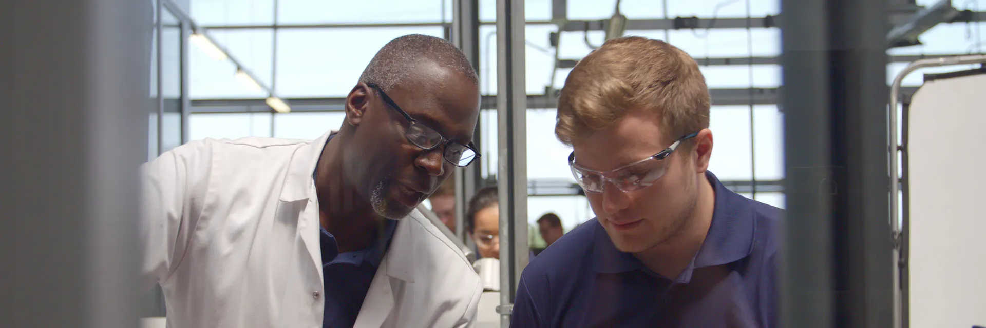 Two men who are engineers, one older on the left who wears a white lab coat, and one younger on the right, who is an apprentice and wears safety glasses, look down at something outside the frame of the image.