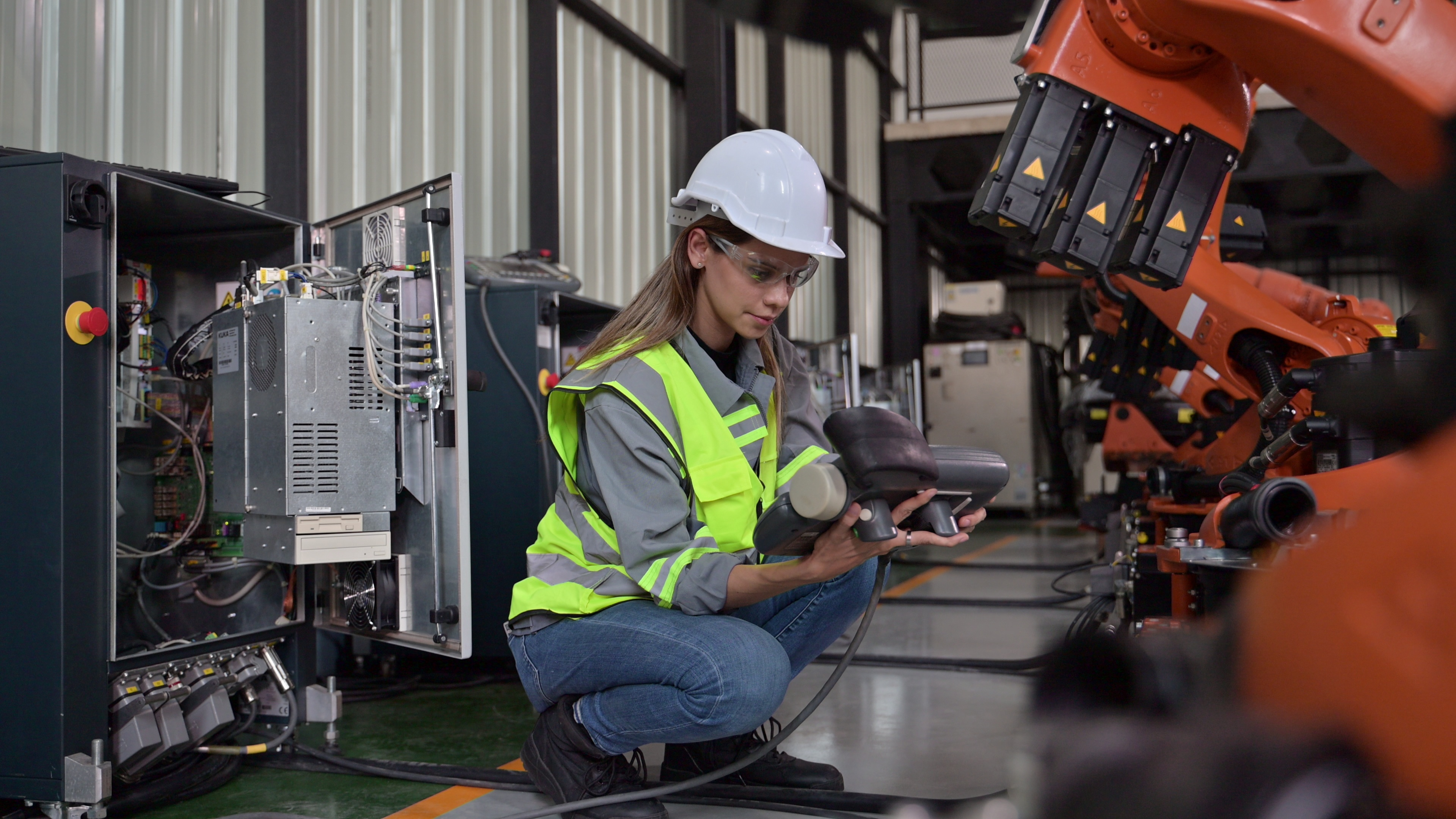 A female maintenance engineer wears a hard hat, safety glasses and a high-visibility vest. She is crouched down and is inspecting a piece of robotic machinery. 