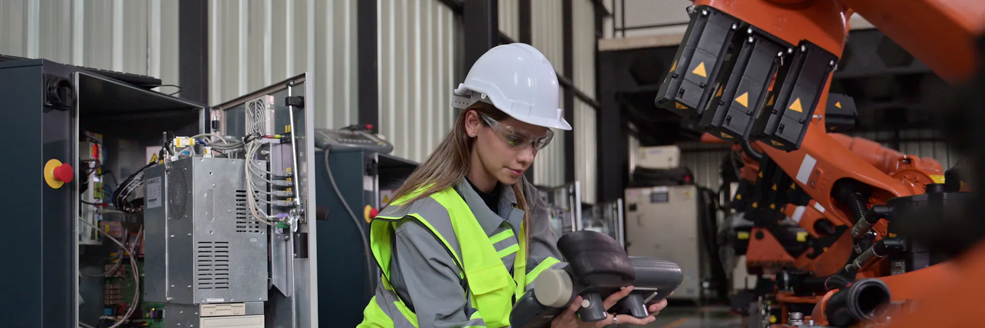 A female maintenance engineer wears a hard hat, safety glasses and a high-visibility vest. She is crouched down and is inspecting a piece of robotic machinery.