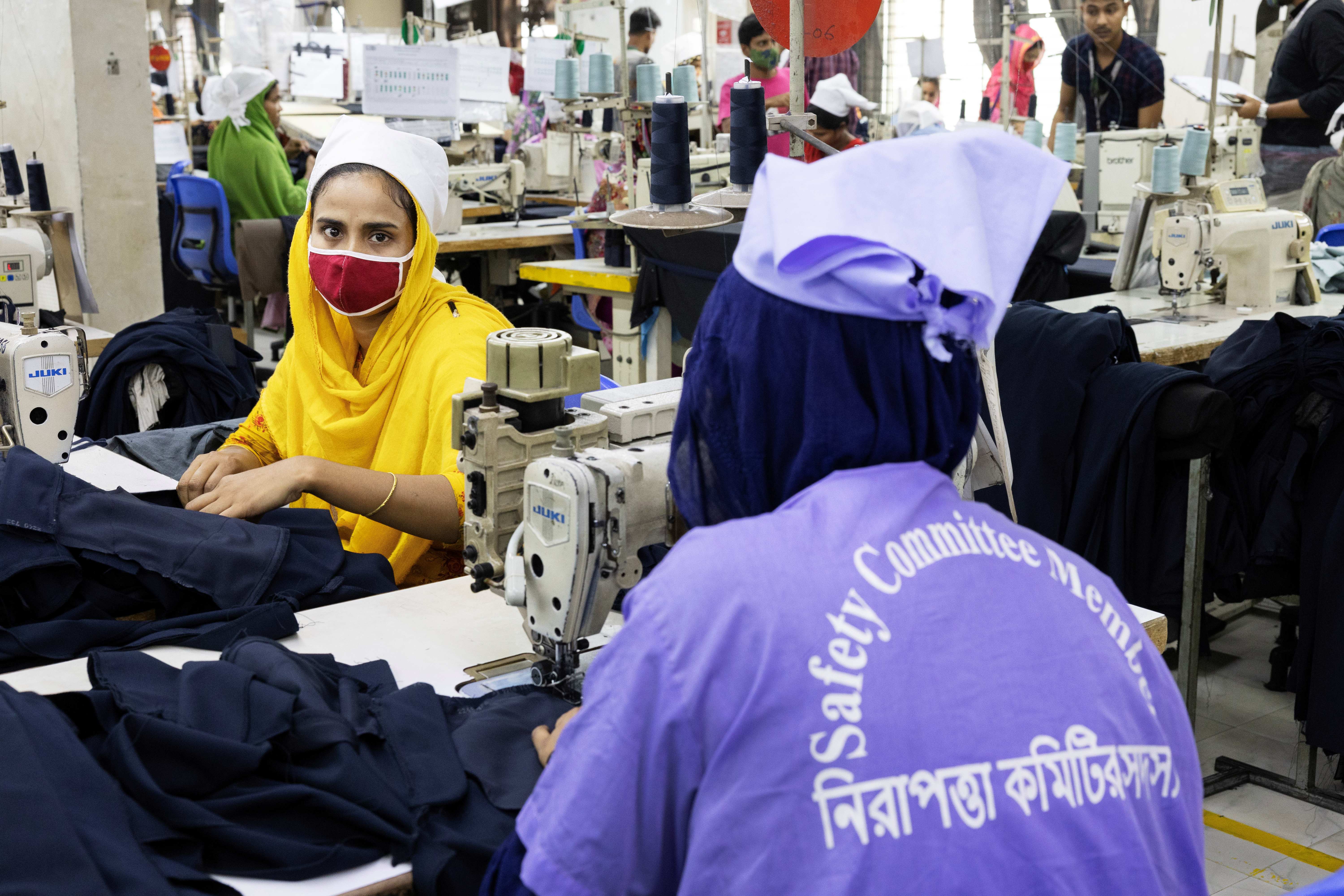 Garment workers operate sewing machines. In the foreground, a female worker wears a purple uniform with "Safety Committee Member" written on the back.  