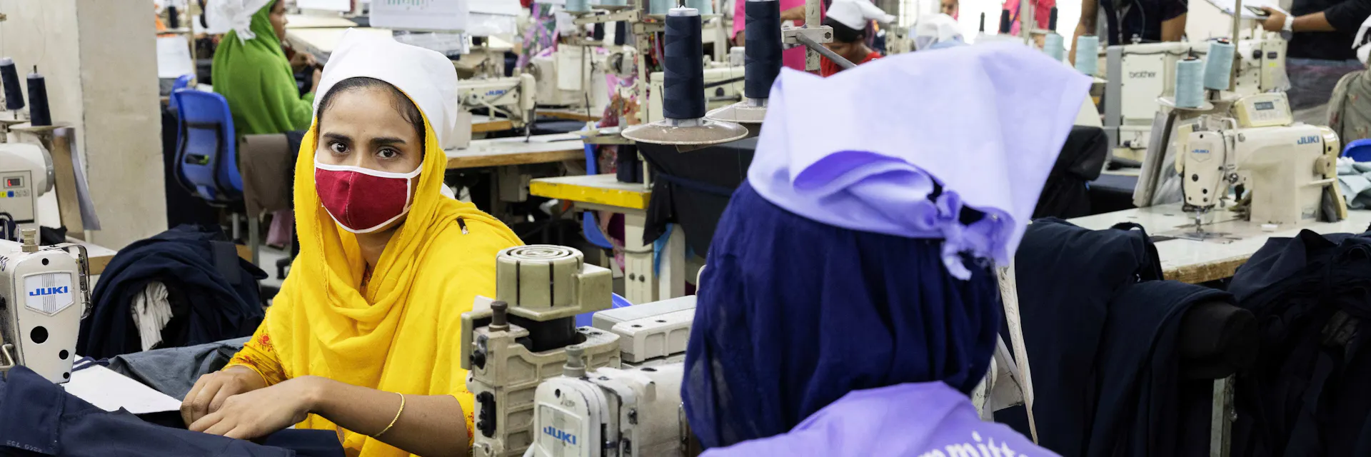 Garment workers operate sewing machines. In the foreground, a female worker wears a purple uniform with "Safety Committee Member" written on the back.