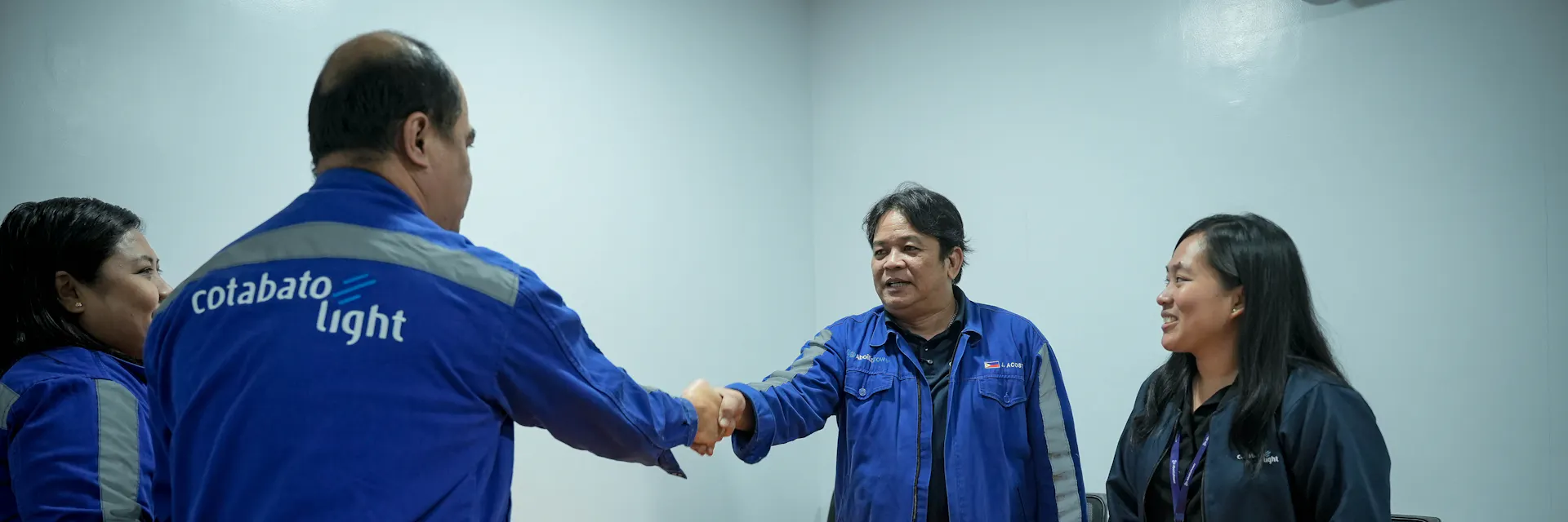 A worker and a manager stand on opposite sides of a table and shake hands, while two other people look on.
