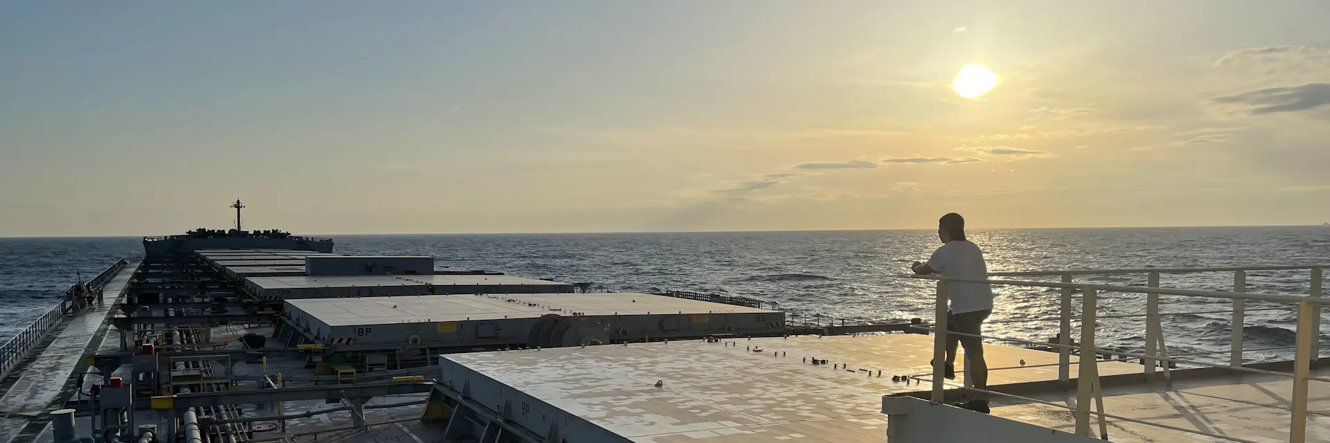 Crew member on the deck of a cargo ship overlooks the ocean at sunset.