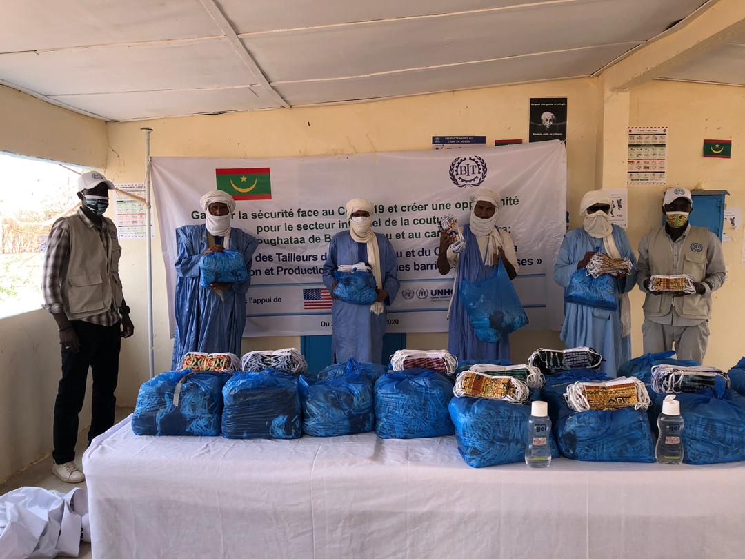 Six hommes qui portent des masques sont debout derrière une table remplie de masques dans des sacs en plastique bleu.  Il y a un panneau derrière eux avec le nom du projet de l’OIT, le logo de l’OIT, le drapeau de la Mauritanie, et le drapeau des Etats-Unis, qui soutient le projet de l’OIT et du UNHCR.
