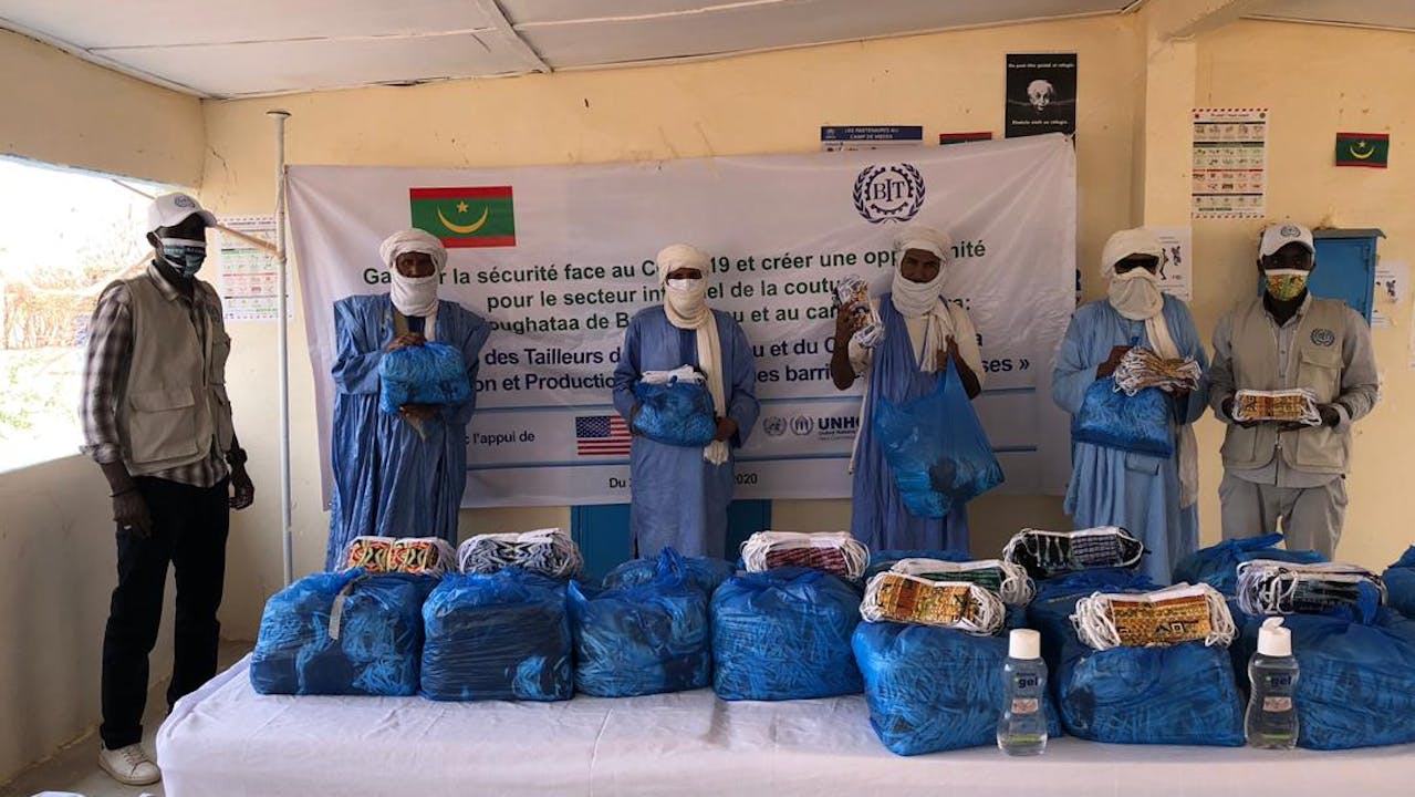 Six men wearing masks stand behind a table. On the table are blue plastic bags filled with masks ready for distribution.  There is a sign behind them with the name of the ILO project, the ILO logo, the flag of Mauritania, and the flag of the United States, which supports the ILO and UNHCR project.