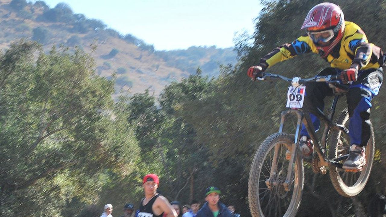 Alfredo Carrasco wears a helmet and protective clothing and rides a mountain bike at a competition while others watch. 