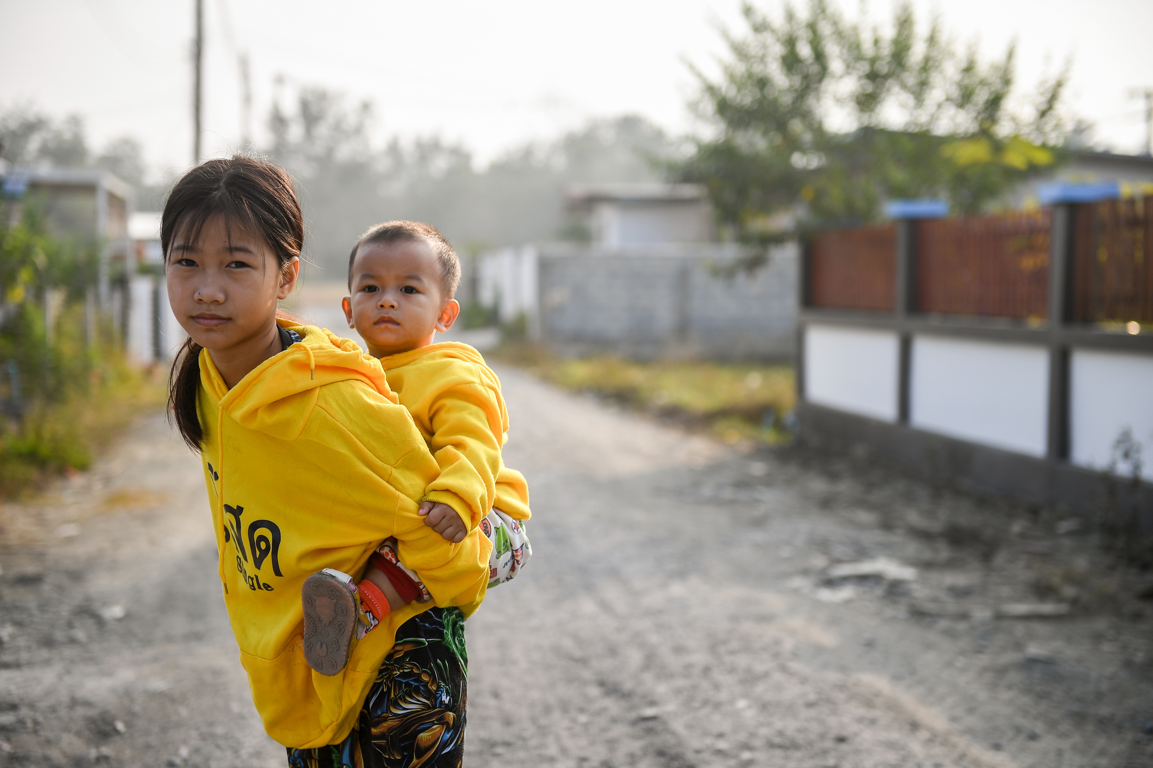Sai Sai's 12-year-old daughter stands on the road and holds her little brother on her back. They both look straight at the camera.