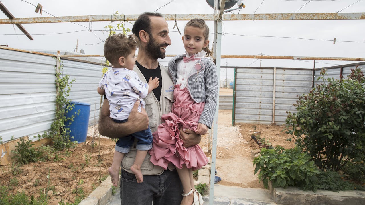 Abdel Halim al Qasir sostiene en brazos a dos de sus hijos, un niño y una niña. Su hija pequeña mira a la cámara. Están en el exterior, hay muros improvisados al fondo y algunas plantas.