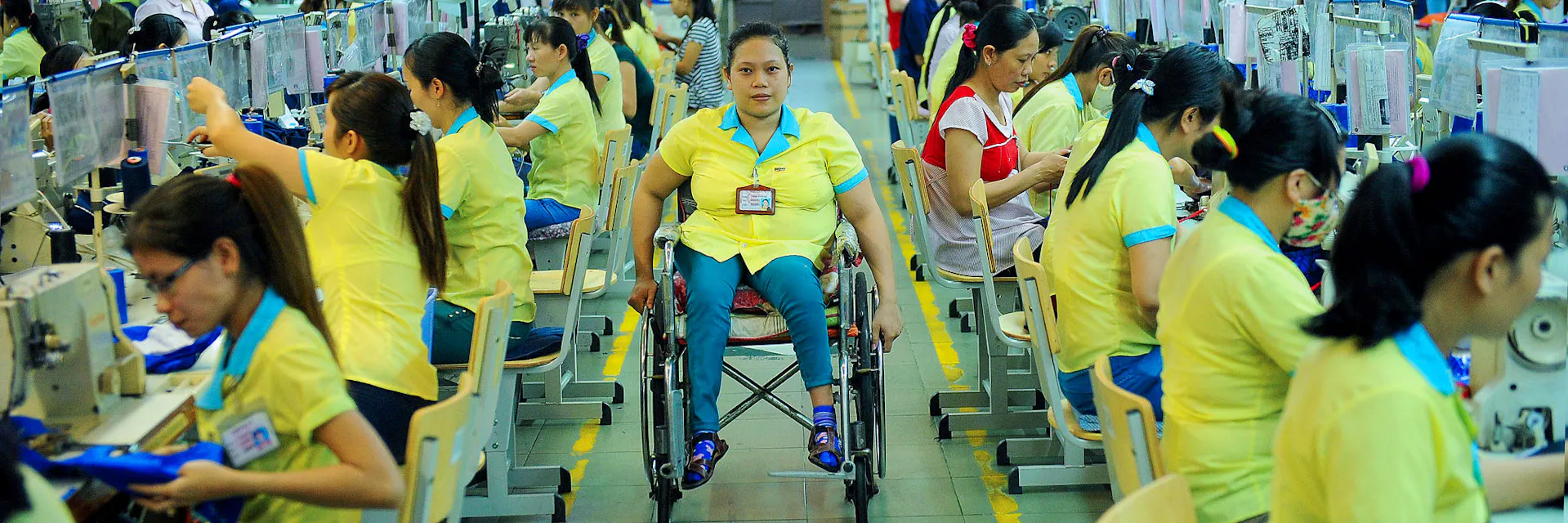 A garment worker in a yellow t-shirt wheels down an aisle in the centre of the photo. She is in a wheelchair. Garment workers all wearing yellow t-shirts work in rows either side of her at sewing machines.