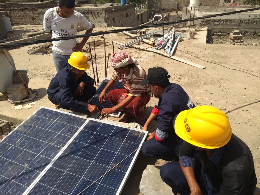 Five men are crouched down next to two solar panels, including Muhammad Taher Muhammad al-Tahri and another apprentice, who wear yellow hard hats.  They are being trained to install solar panels.   