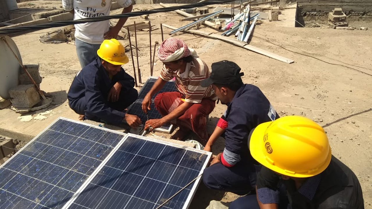 Cinco hombres están agachados junto a dos paneles solares, entre ellos Muhammad Taher Muhammad al-Tahri y otro aprendiz, que llevan cascos amarillos. Están recibiendo formación para instalar paneles solares.   