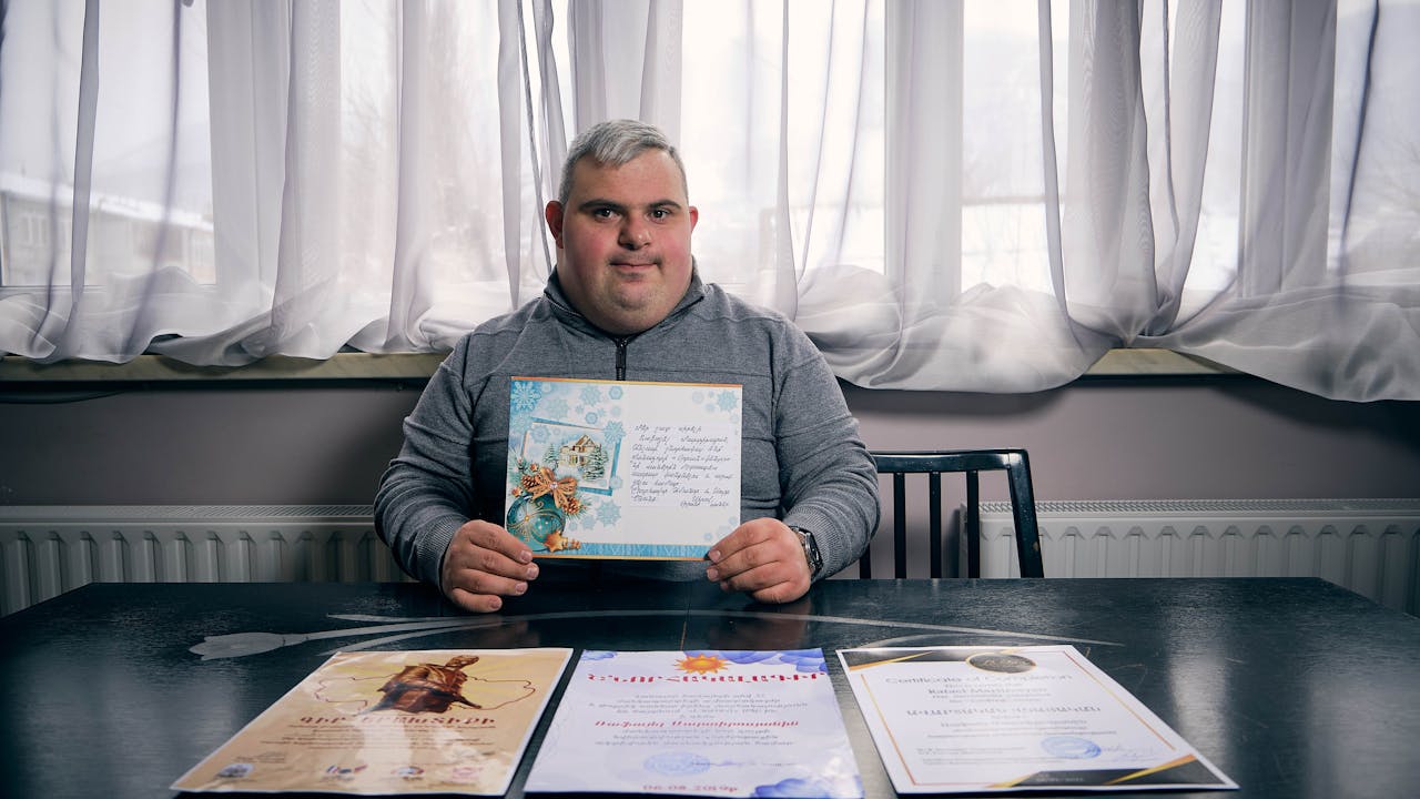 Rafael Martirosyan sits on a chair and holds up one of his certificates for his volunteer work. Other certificates are laid out on the table in front of him. 