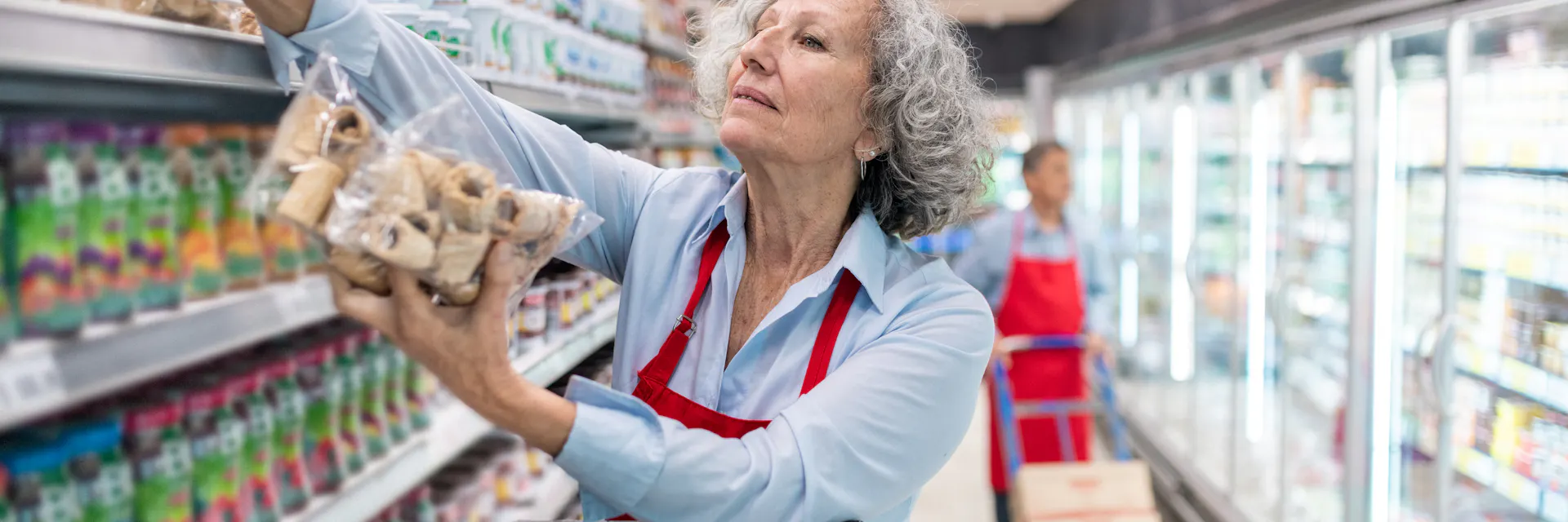 Personne d'âge mûr travaillant dans un supermarché. Au premier plan, une femme aux cheveux grisonnants range des marchandises dans les rayons du magasin.