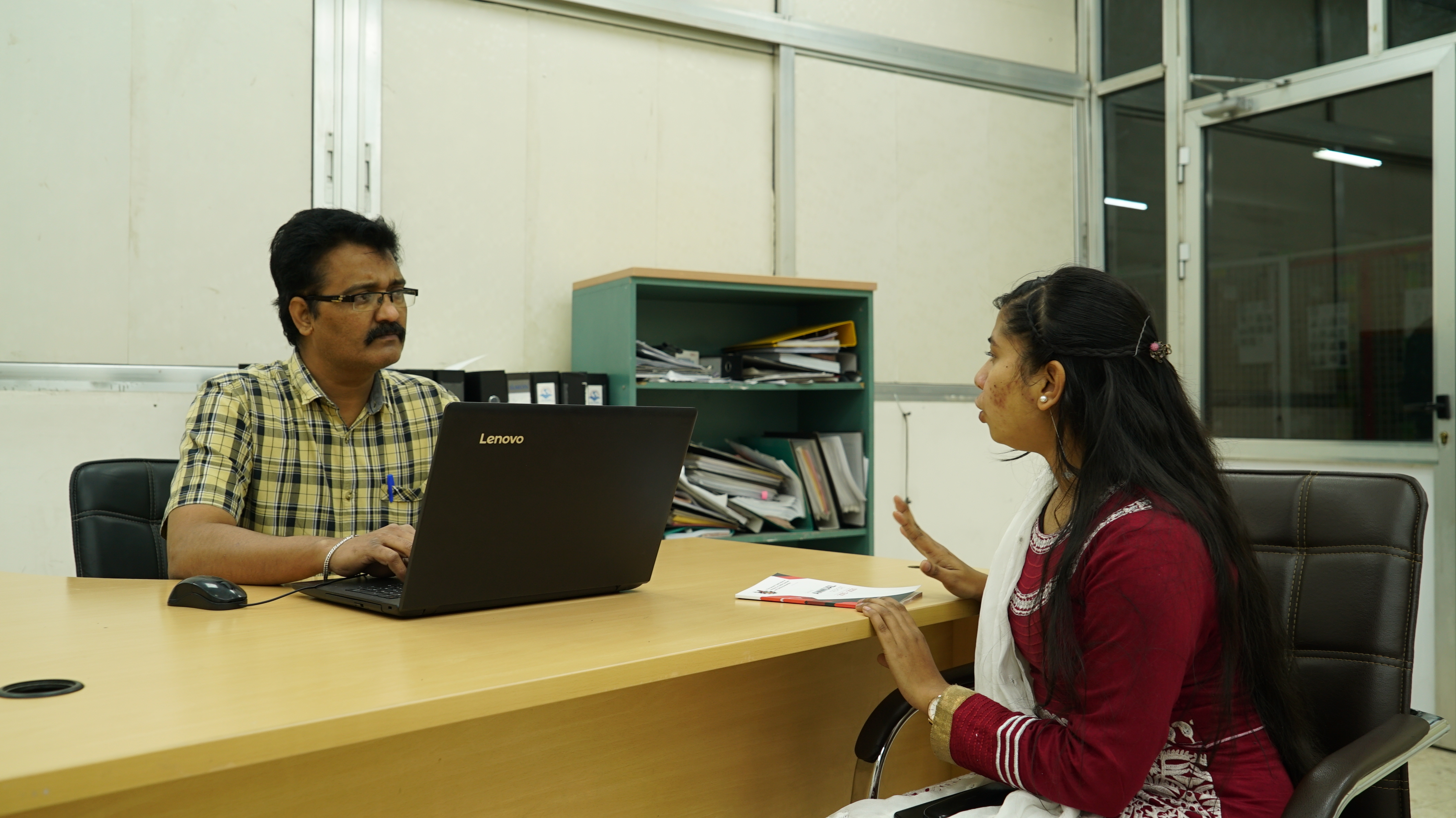 Maya Aktar sits opposite and discusses with a garment factory manager who is sitting at his desk.