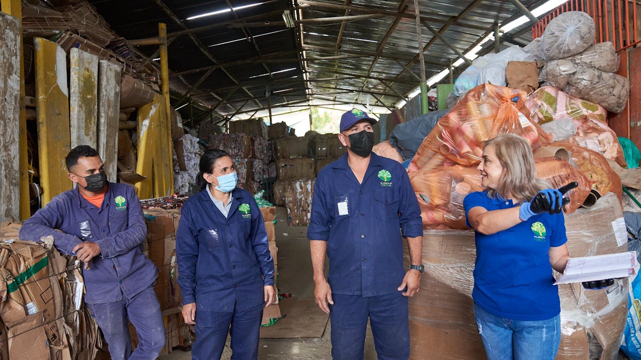 Martha Elena Iglesias holds a clipboard and points at something in the recycling warehouse.  Three waste pickers look on.