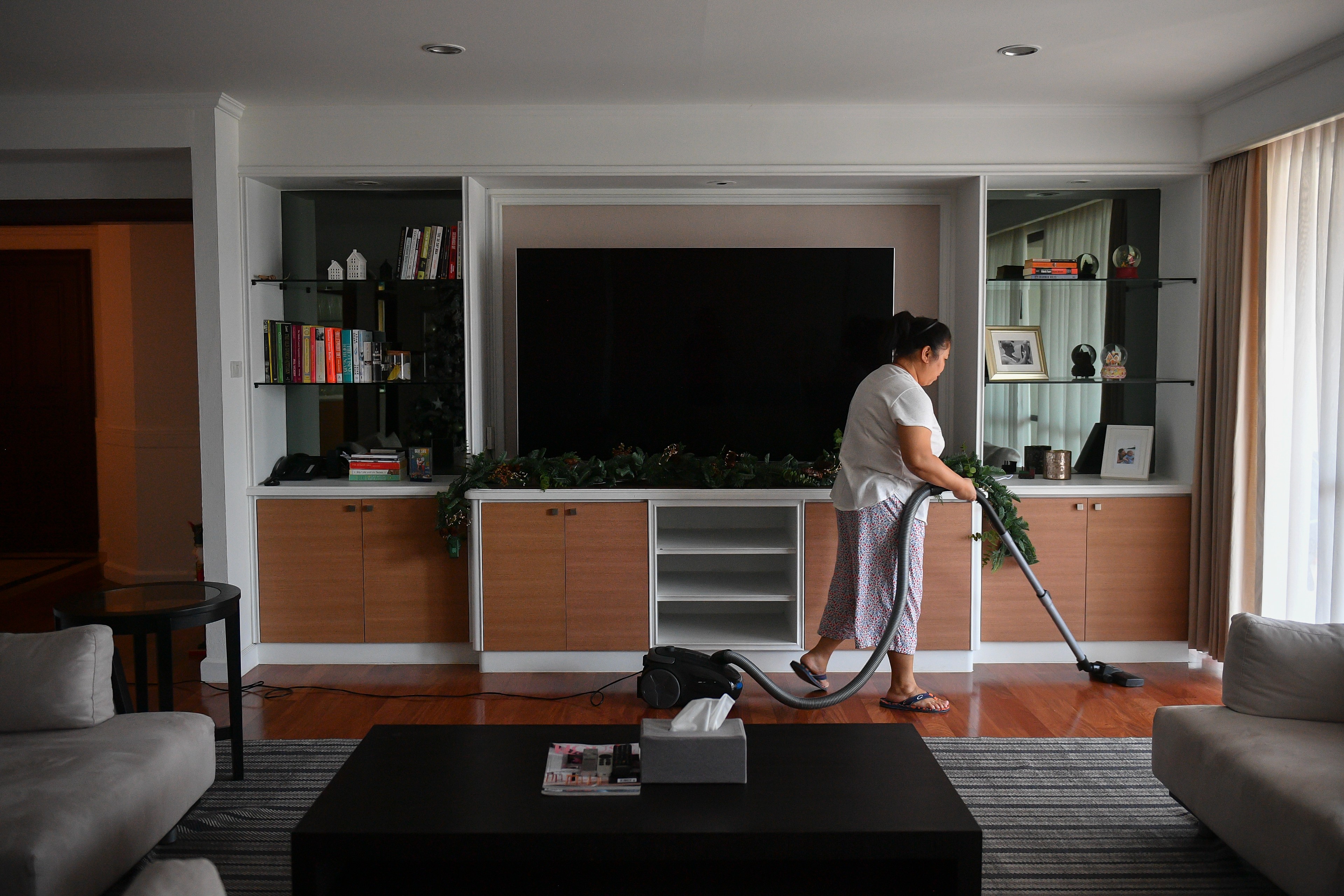 Champa passe l'aspirateur dans le salon de son employeur, avec une télévision à grand écran en arrière-plan.