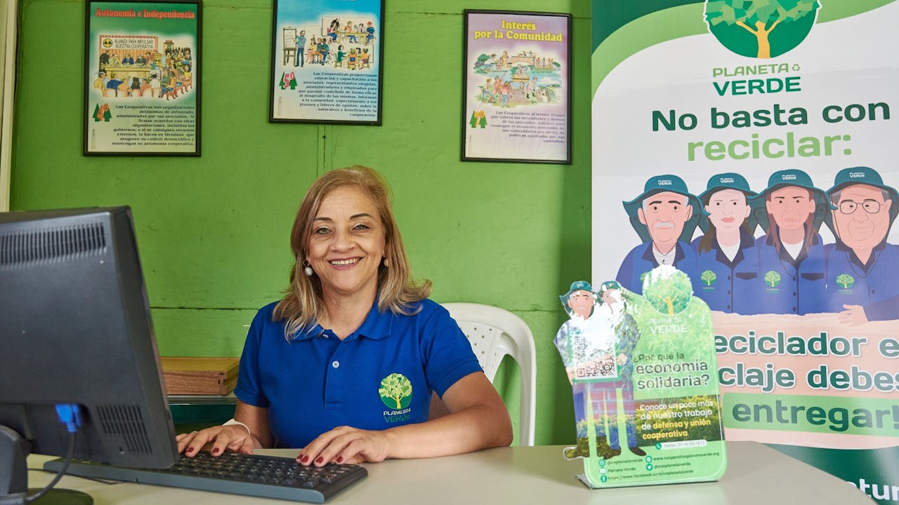 Martha Elena Iglesias se sienta en su escritorio y utiliza un ordenador.  A su lado hay una pancarta de Planeta Verde. 