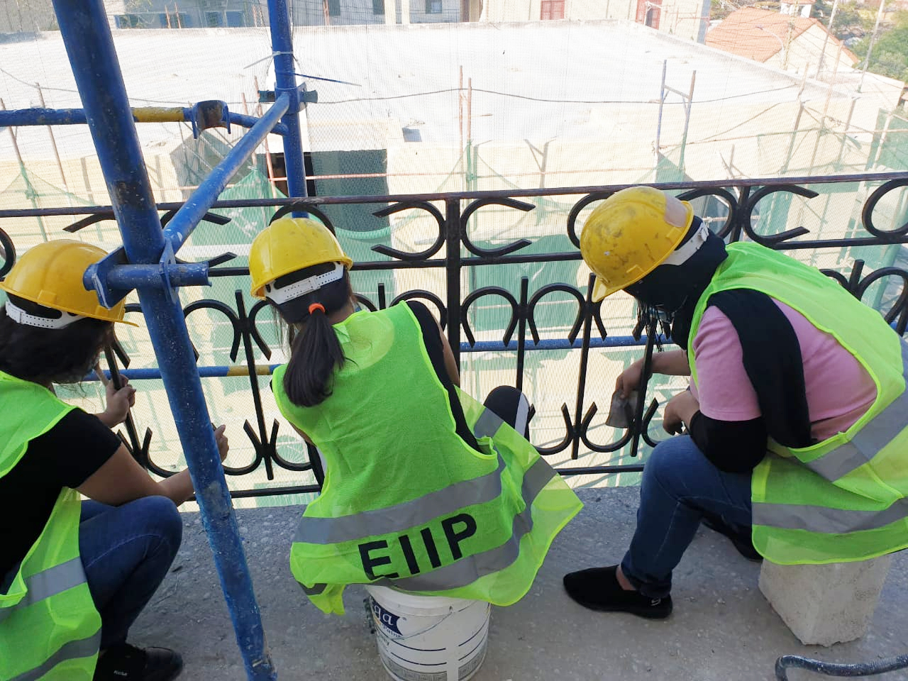 Trabajadoras reparan una balaustrada metálica. Llevan cascos amarillos y camisetas fluorescentes con la inscripción EIIP, que significa Programa de Inversión Intensiva en Empleo de la OIT.