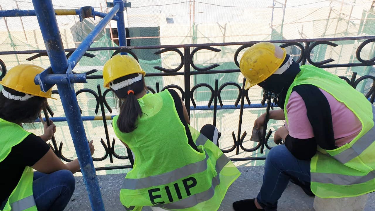 Female workers repair metal balustrade. They wear yellow hard hats and fluorescent jerseys with EIIP written on them, which stands for the ILO’s Employment-Intensive Investment Programme.