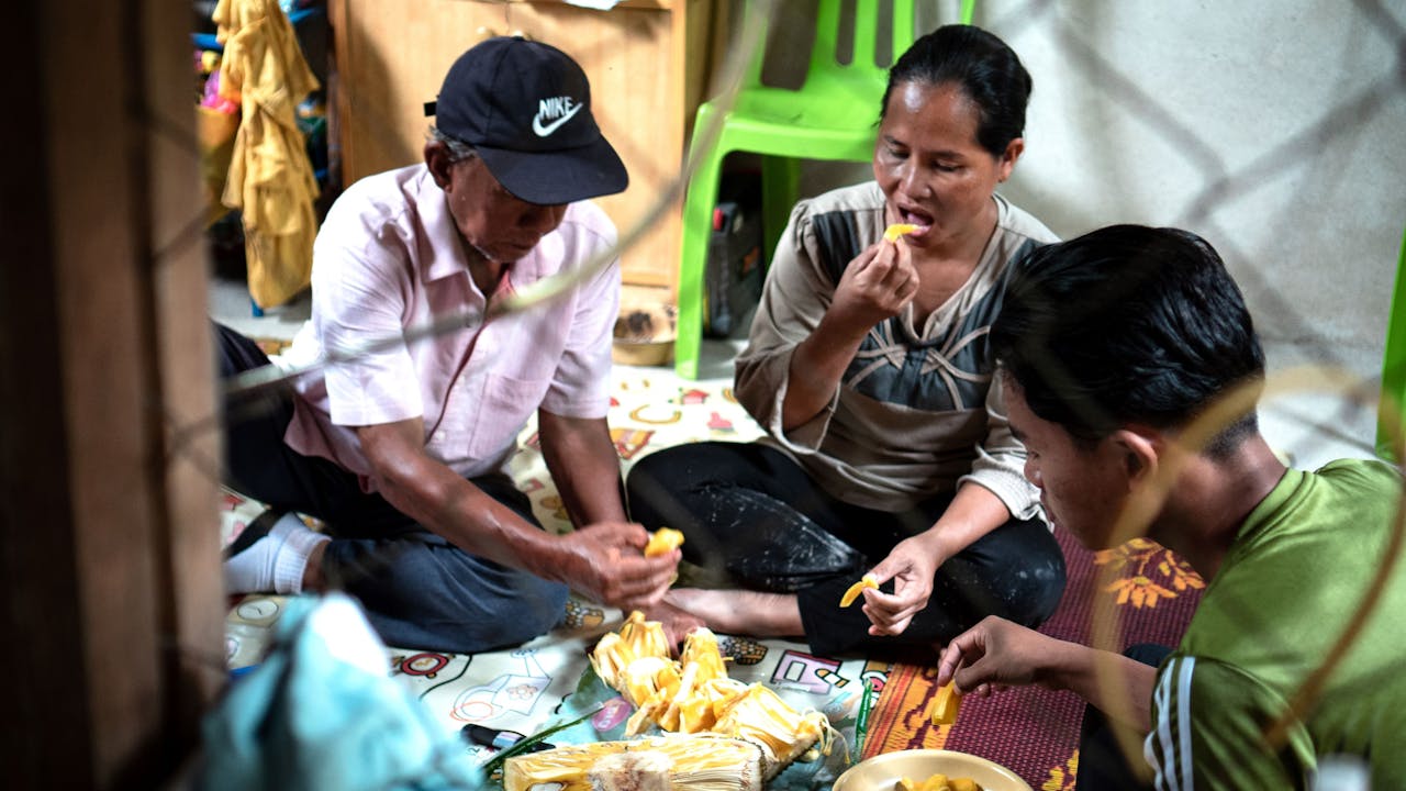 Maimun sits cross legged in her home and eats jackfruit with her husband and one of her teenage sons.