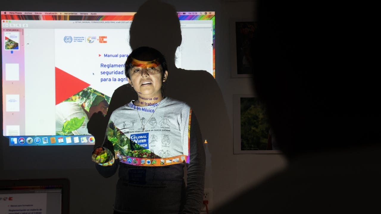 Briseida Venegas Ramos stands in front of a screen which shows ILO safety and health messages.