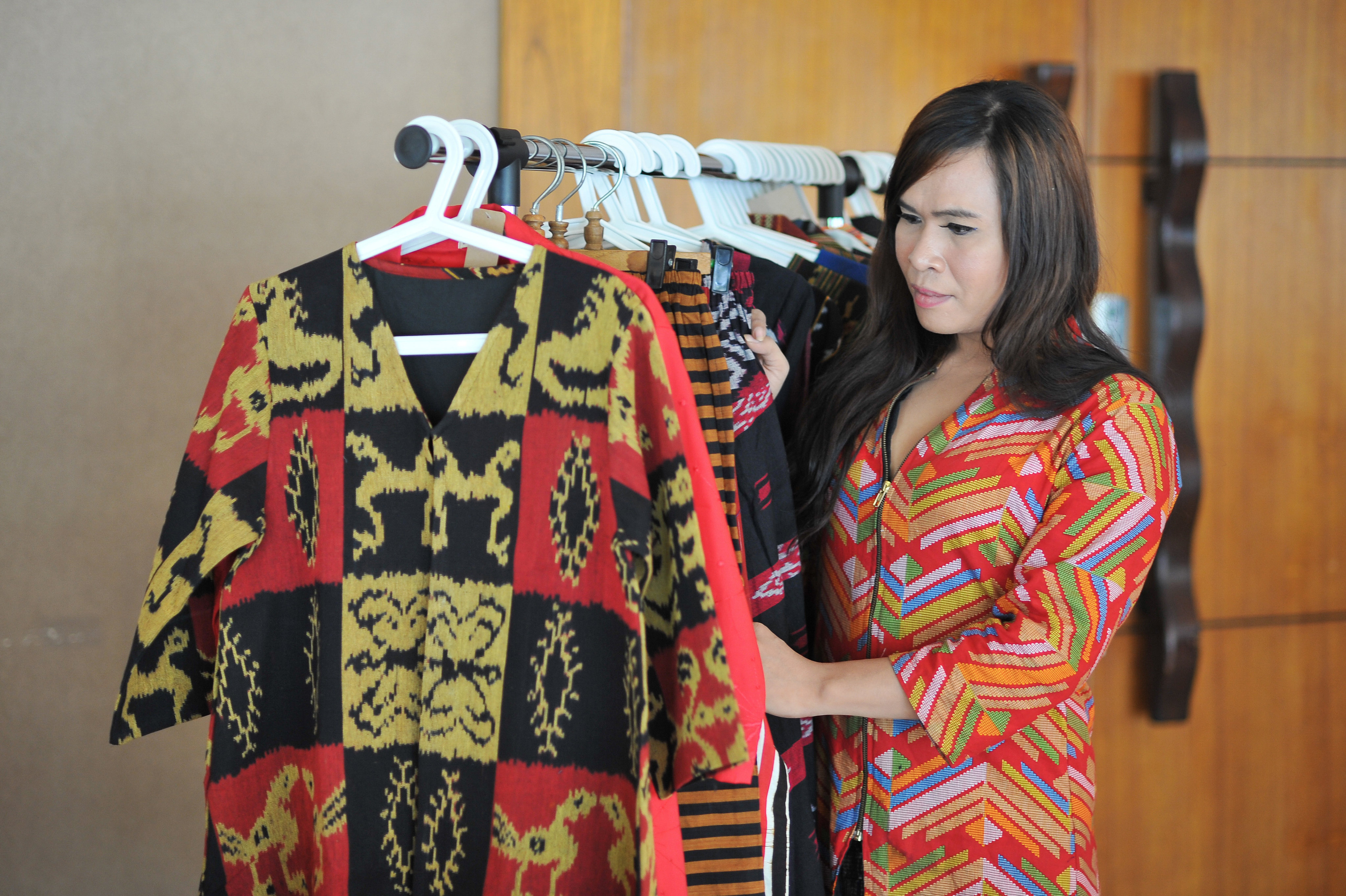 A transwoman, Emi, examines a row of clothing on a rack.  She wears a brightly coloured outfit. Emi works in marketing in Indonesia at a garment business called Sri Kendes, which is managed by a transgender community.