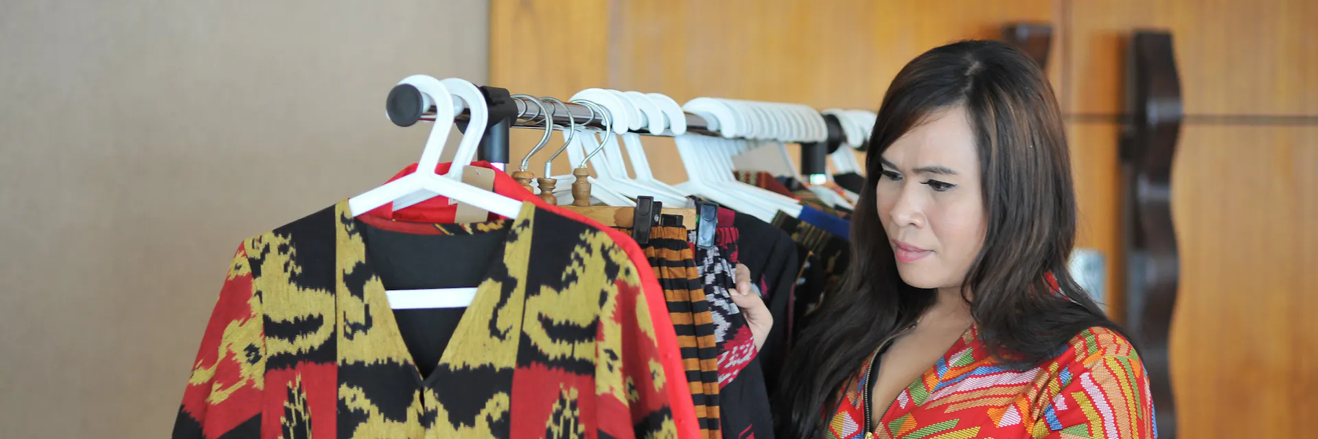 A transwoman, Emi, examines a row of clothing on a rack. She wears a brightly coloured outfit. Emi works in marketing in Indonesia at a garment business called Sri Kendes, which is managed by a transgender community.