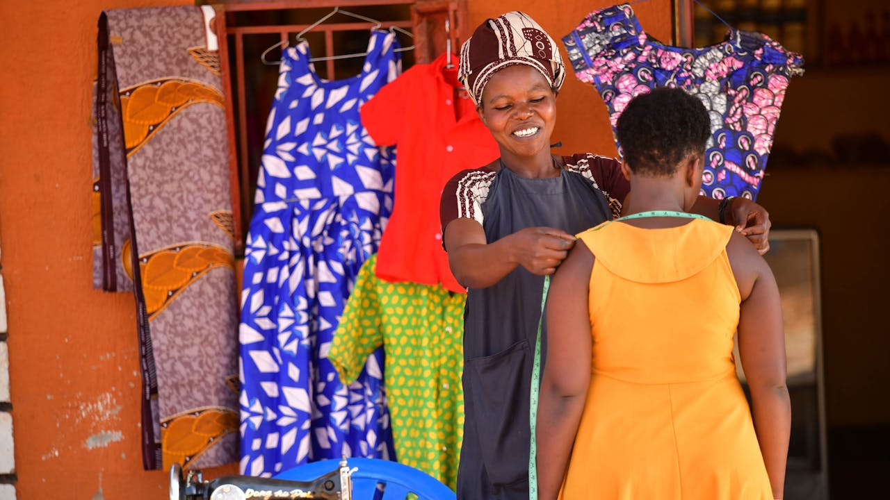Angelique Kahindo, a tailor, measures a customer who wears a yellow dress.  In front of them is a sewing machine. In the background is a bright orange wall and a window frame with fabric hanging around it.  