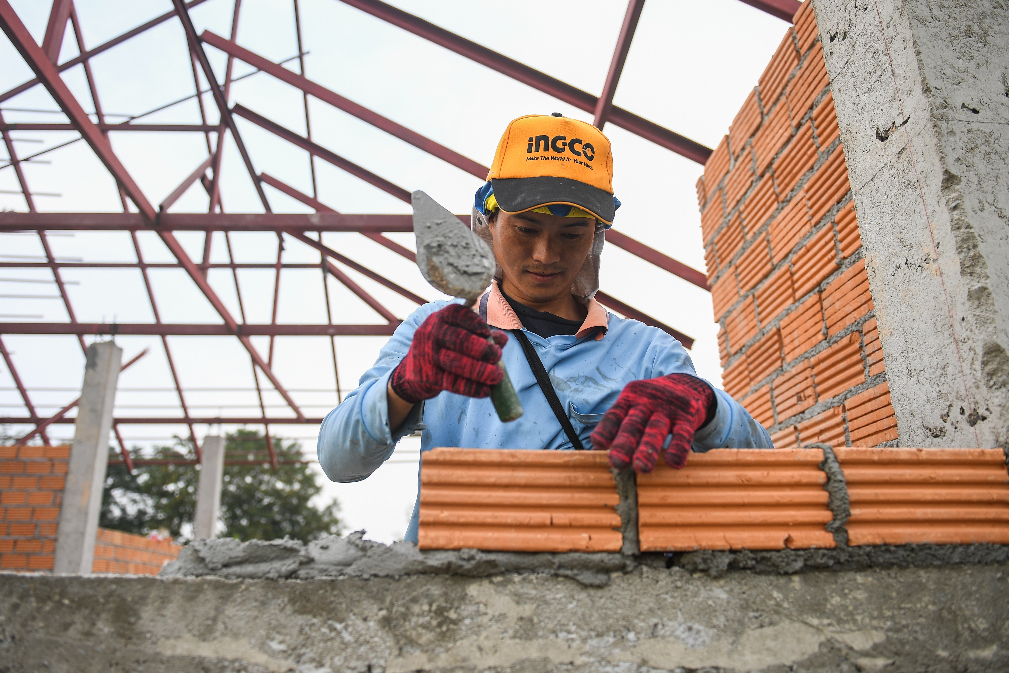 Sai Sai lays bricks at a construction site. Metal roof structure is in the background. 