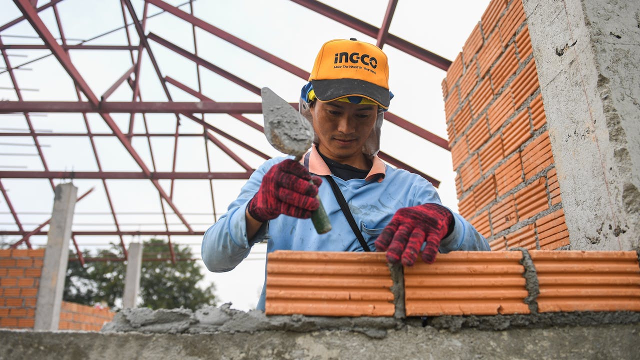 Sai Sai lays bricks at a construction site. Metal roof structure is in the background. 
