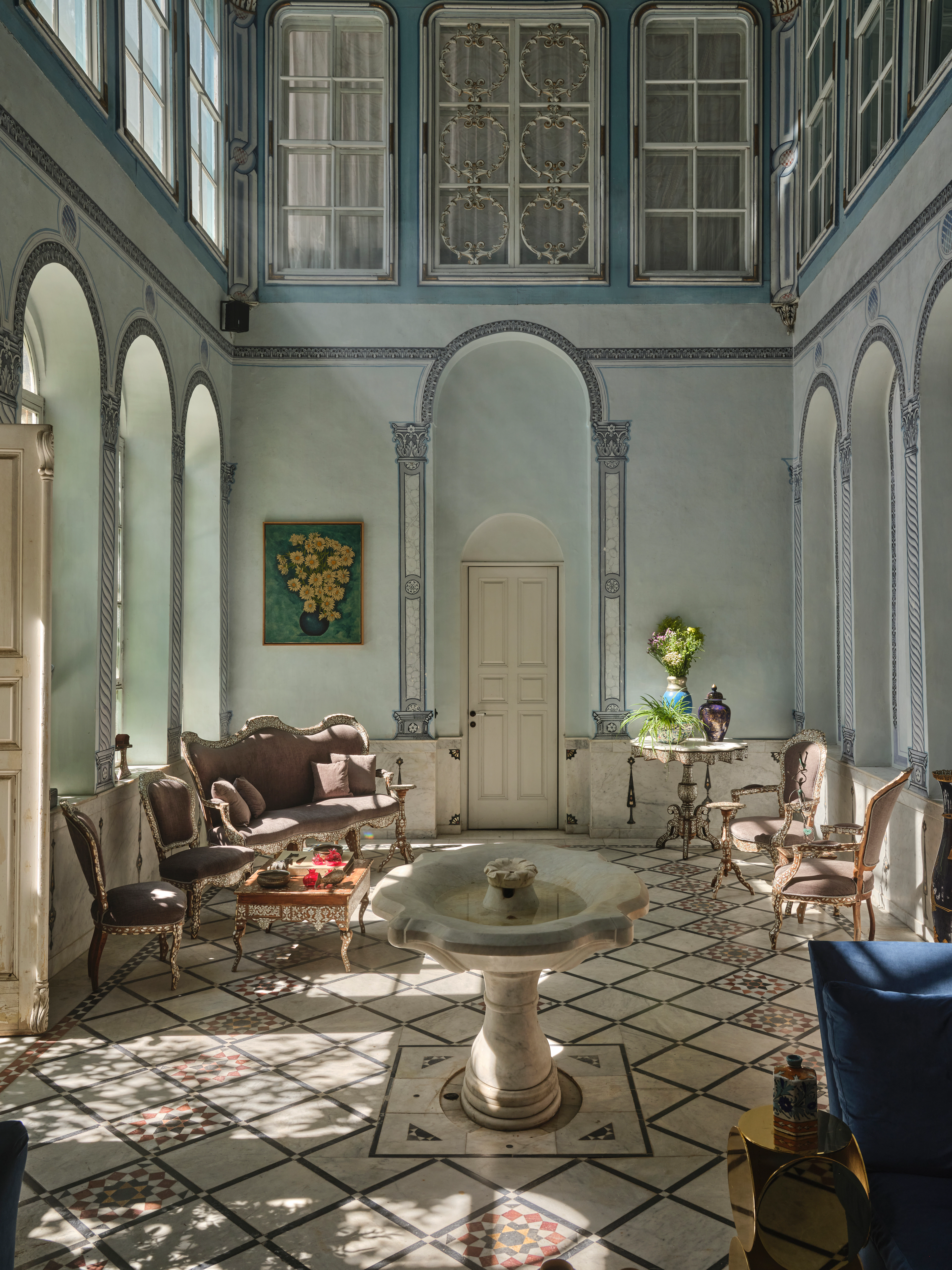 Blue and white sitting room with a central stone basin table, patterned floor, tall windows, and classic upholstered furniture.