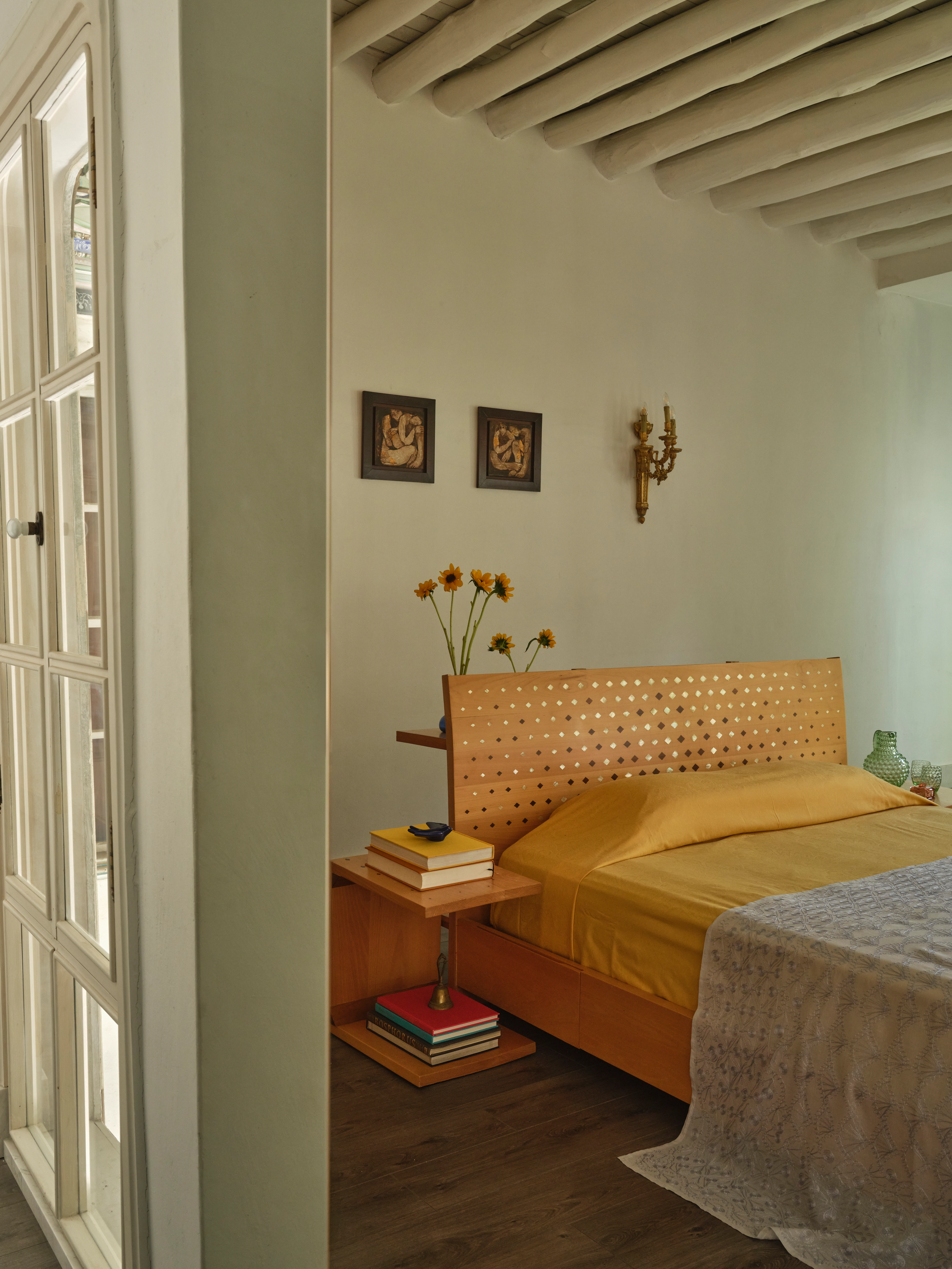 Sunlit bedroom with a perforated wooden headboard, mustard bedding, framed artwork, and exposed ceiling beams.