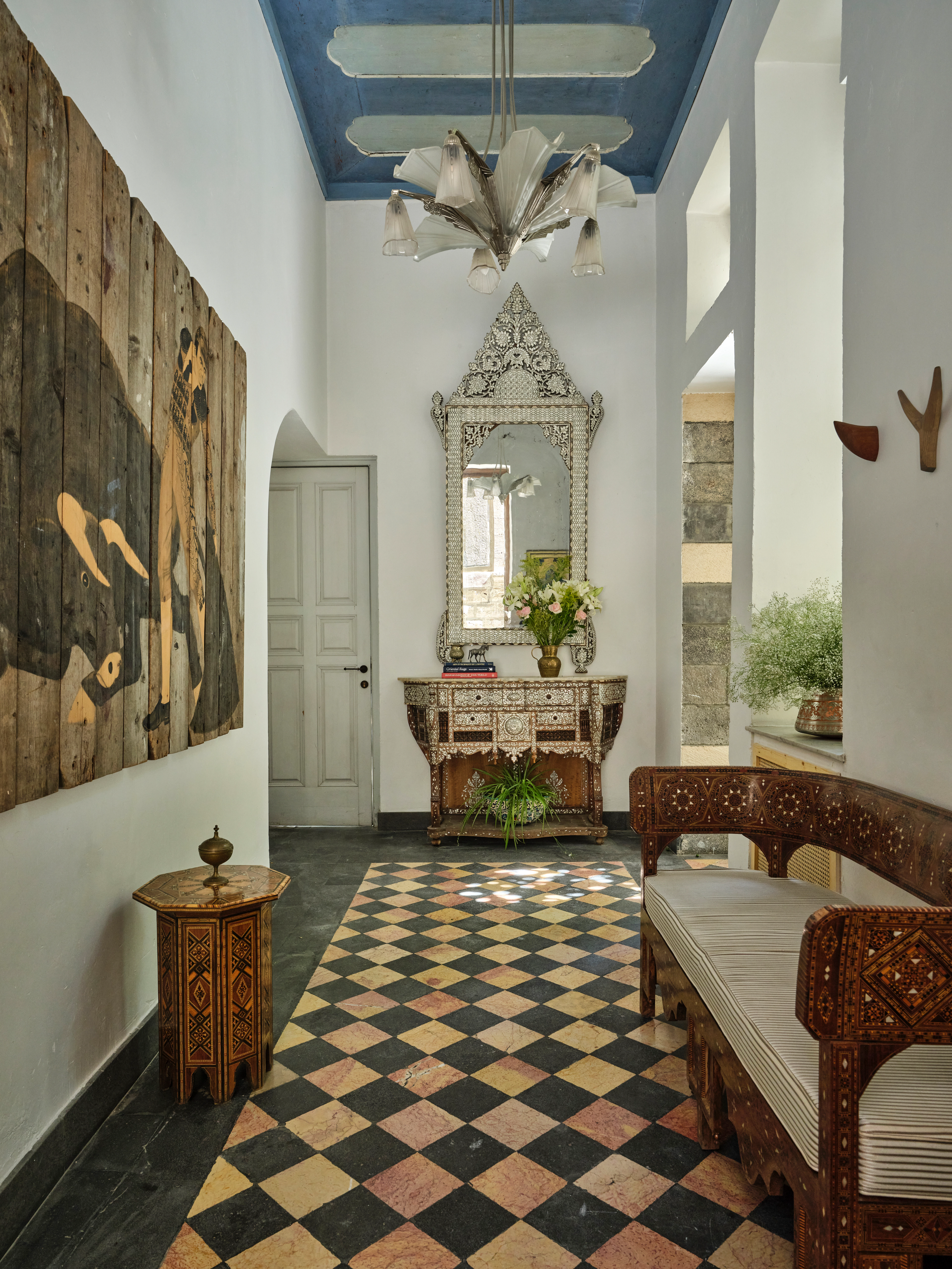 Interior hallway with a blue painted ceiling, sculptural chandelier, ornate mirror and console, carved seating, artwork, and patterned floor.