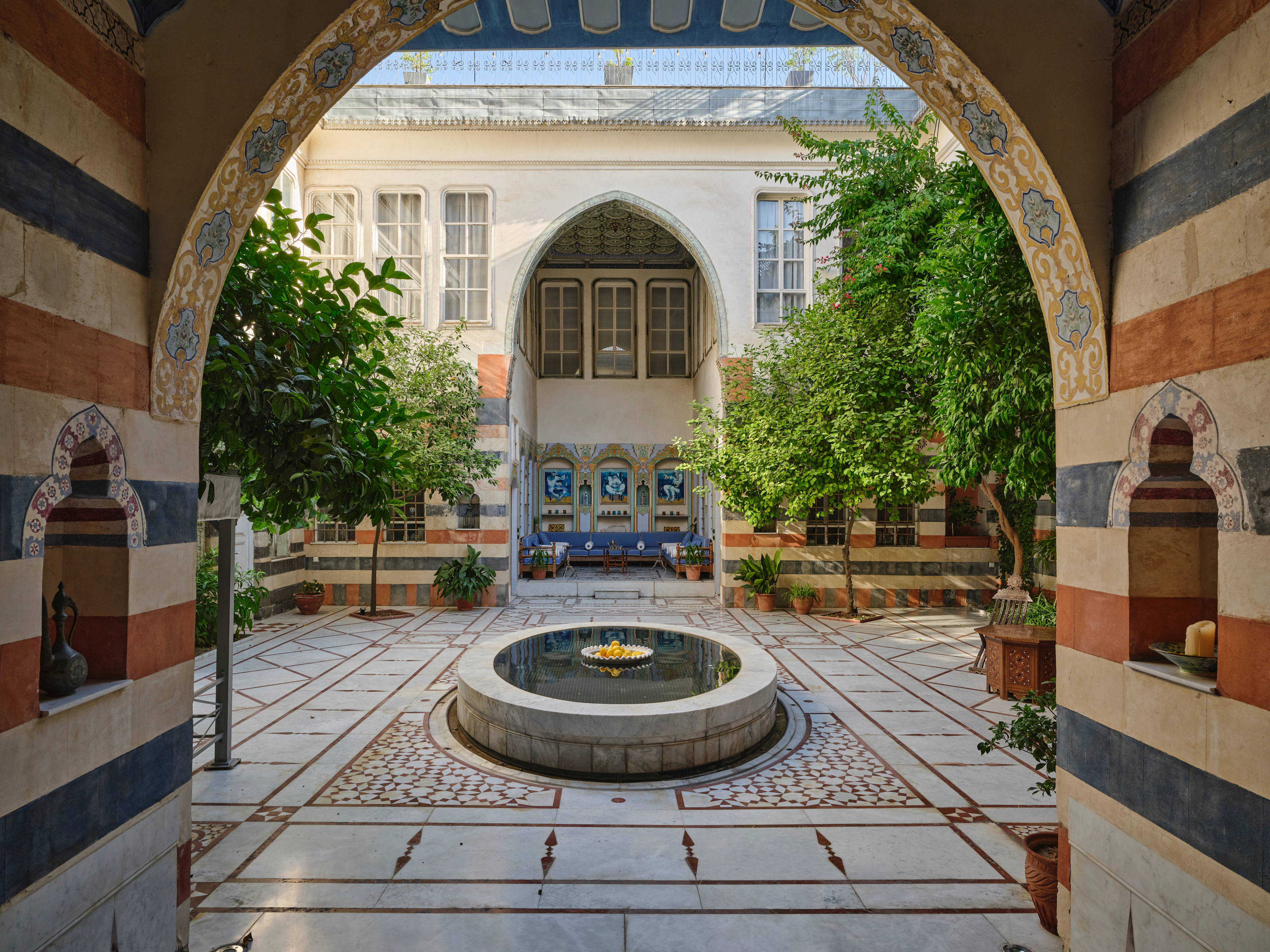 Historic Damascus courtyard framed by a painted arch, with a central fountain, patterned marble floor, citrus trees, and a blue seating area.
