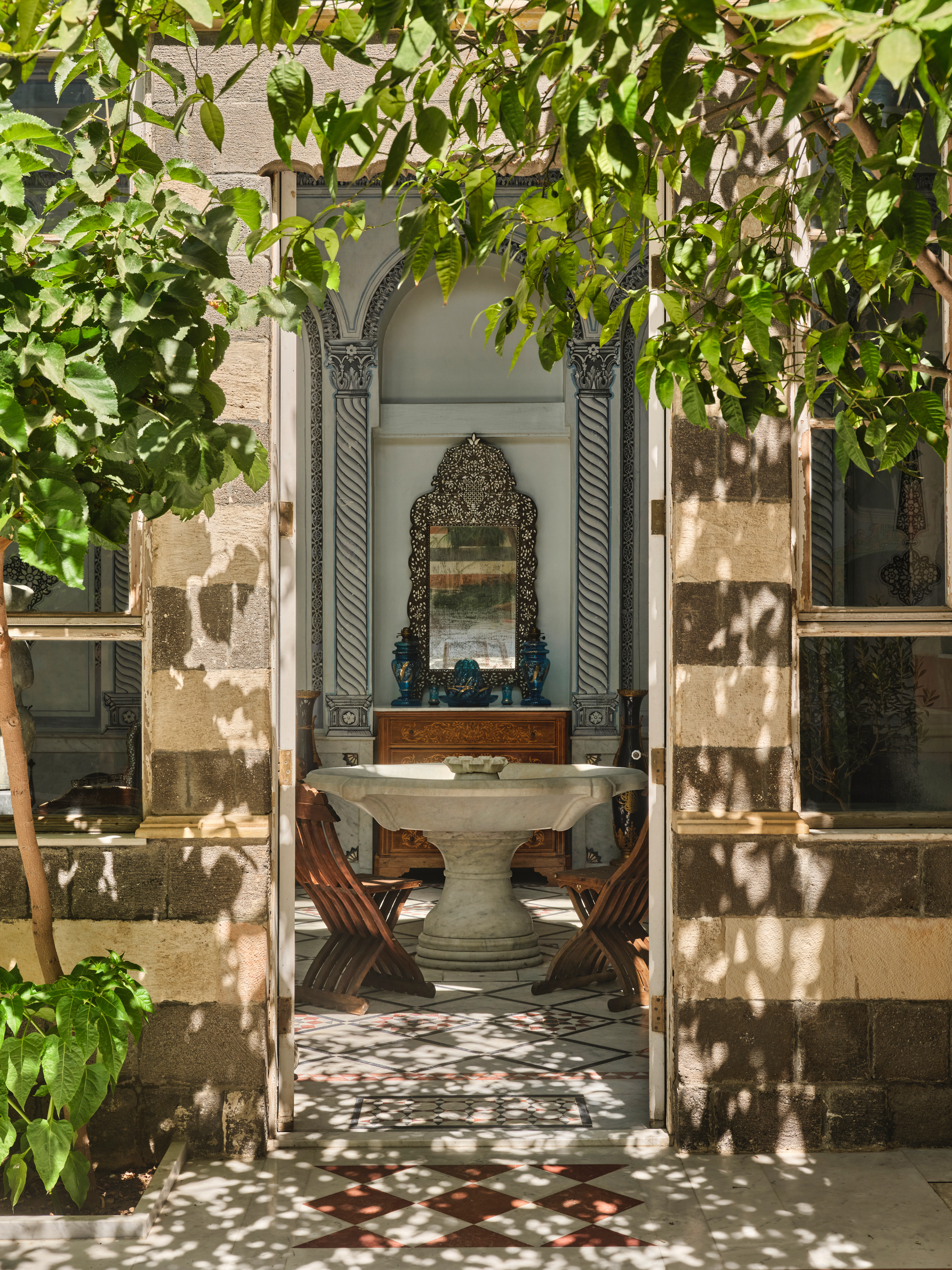 View through a doorway into an ornate blue and white room with a stone table, arched wall details, and leafy branches.