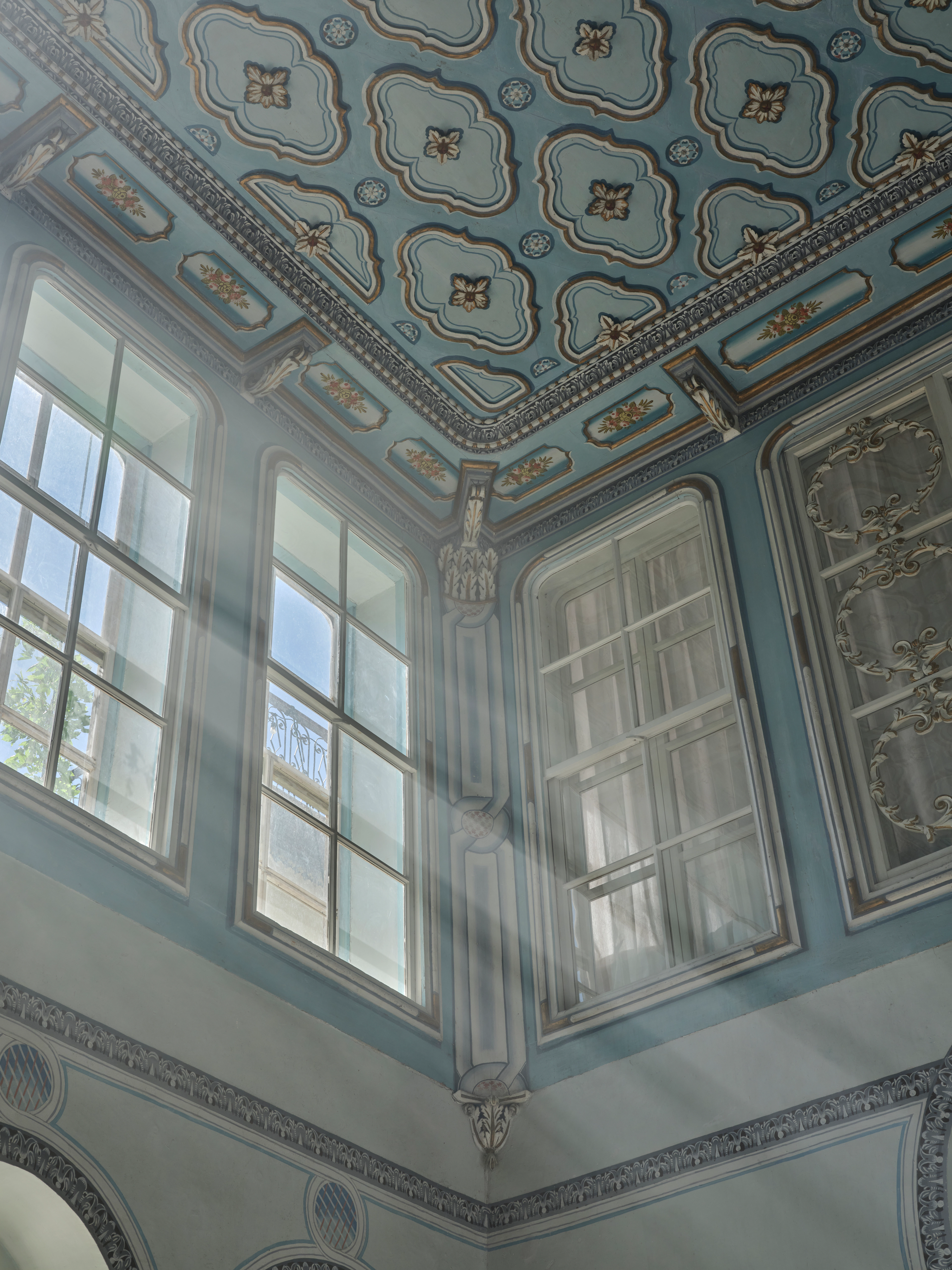 Close view of an ornate blue painted ceiling and tall windows in a light-filled room.