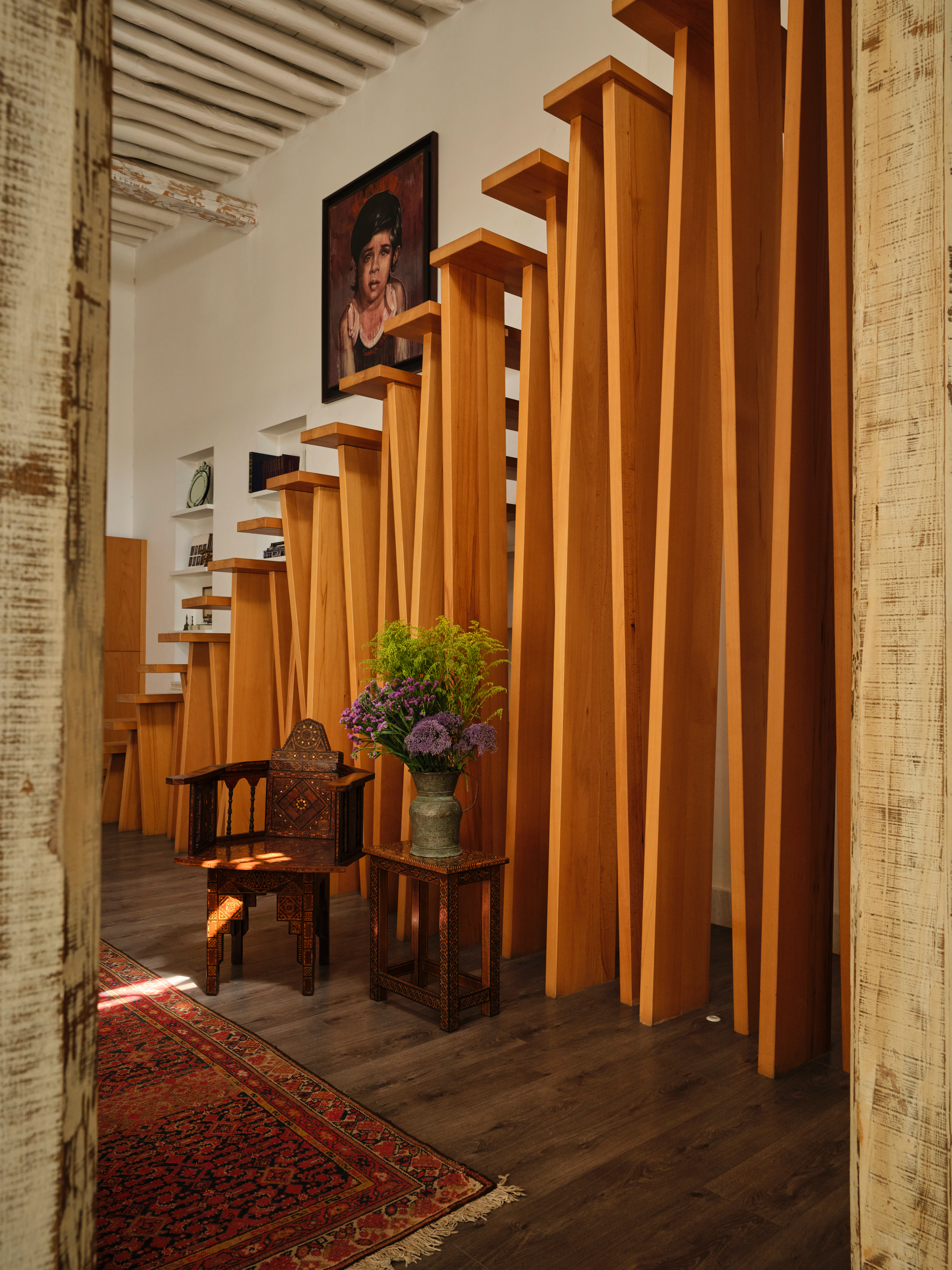 Interior view of a timber staircase with sculptural vertical supports, a carved chair, artwork, and patterned rugs.