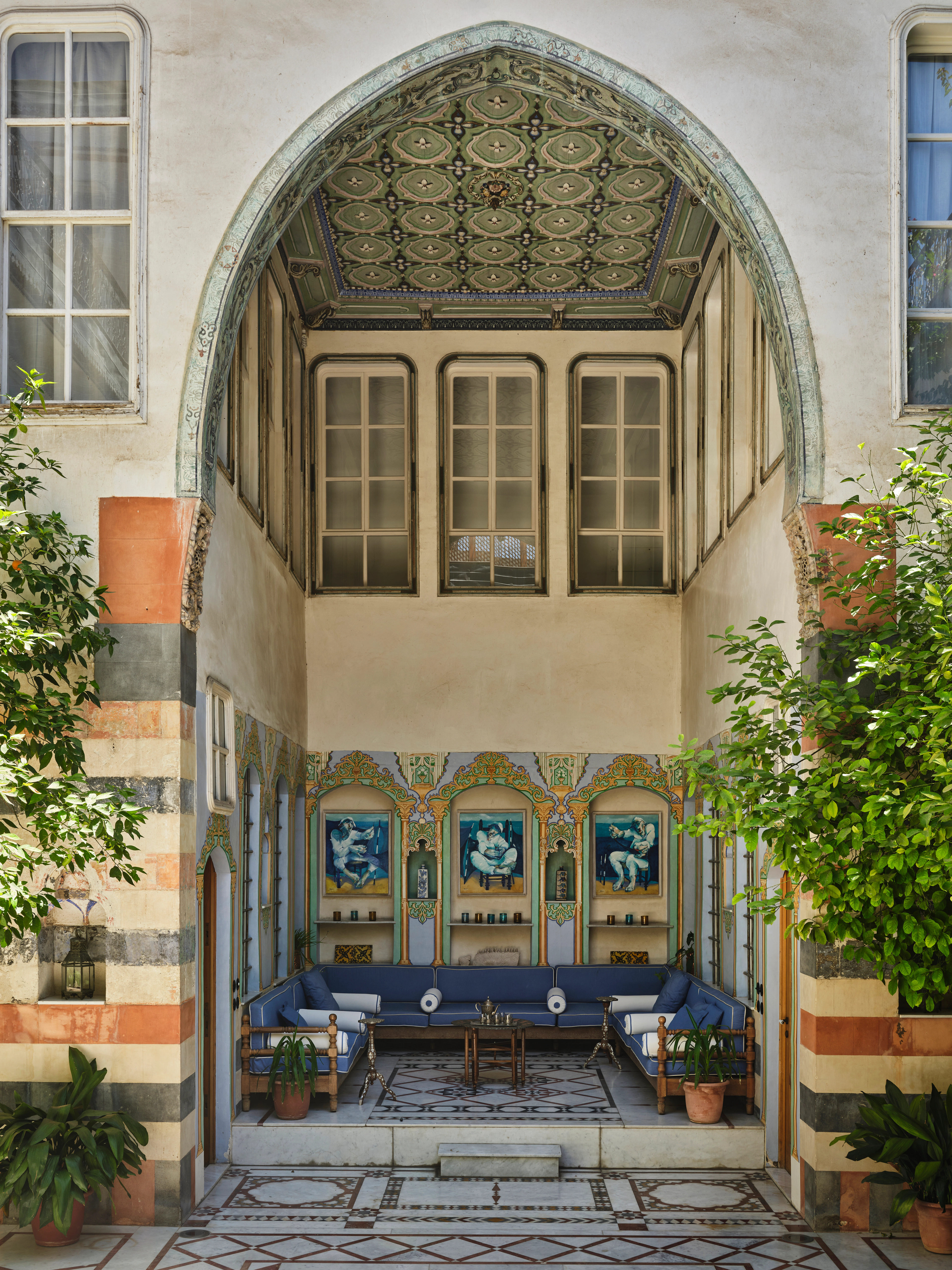 Ornate courtyard seating area with blue sofas, patterned tile floors, painted wall panels, and lush plants.