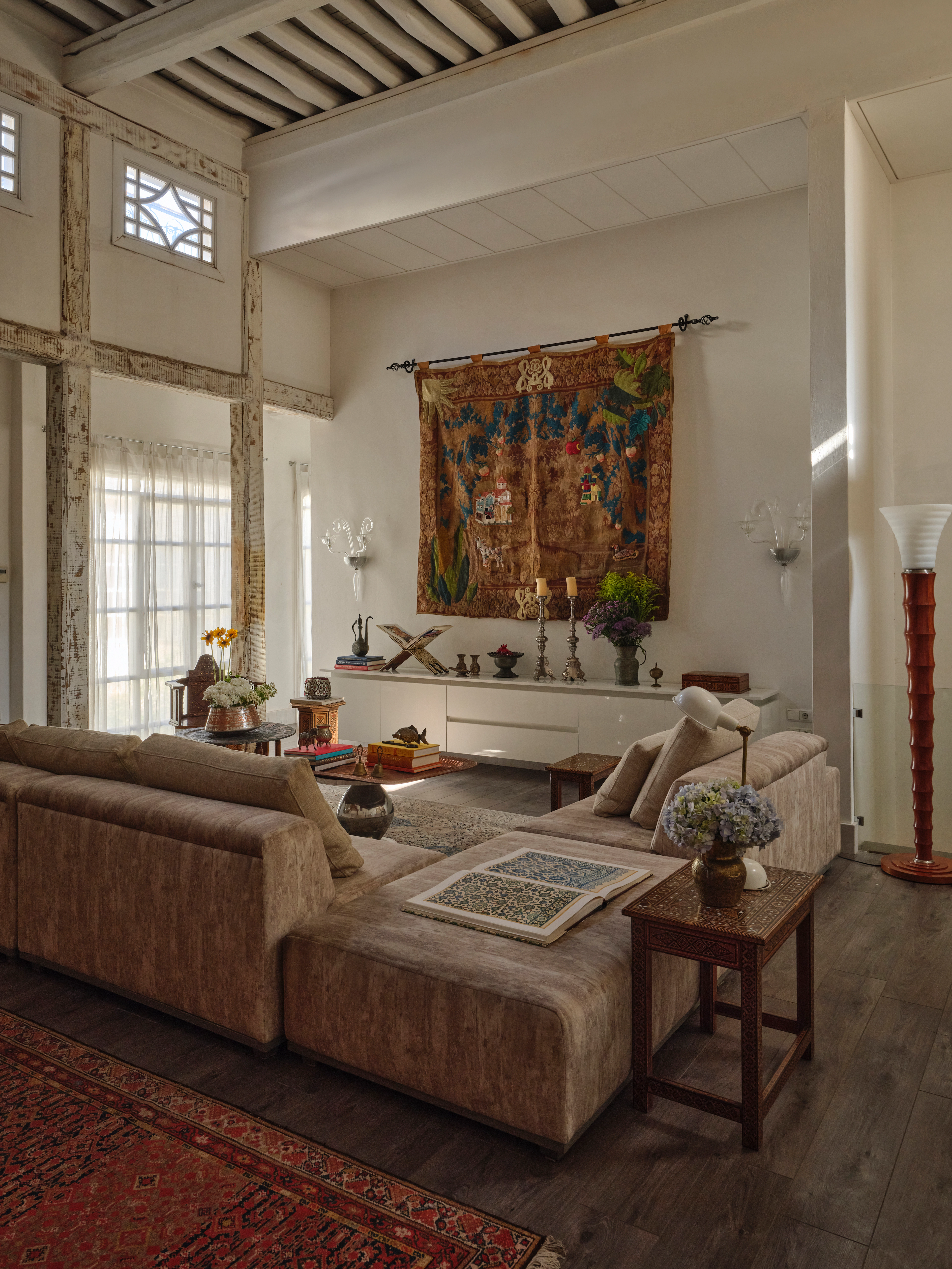 Living room with linen sofas, exposed timber beams, a large colorful wall hanging, and layered Persian rugs.