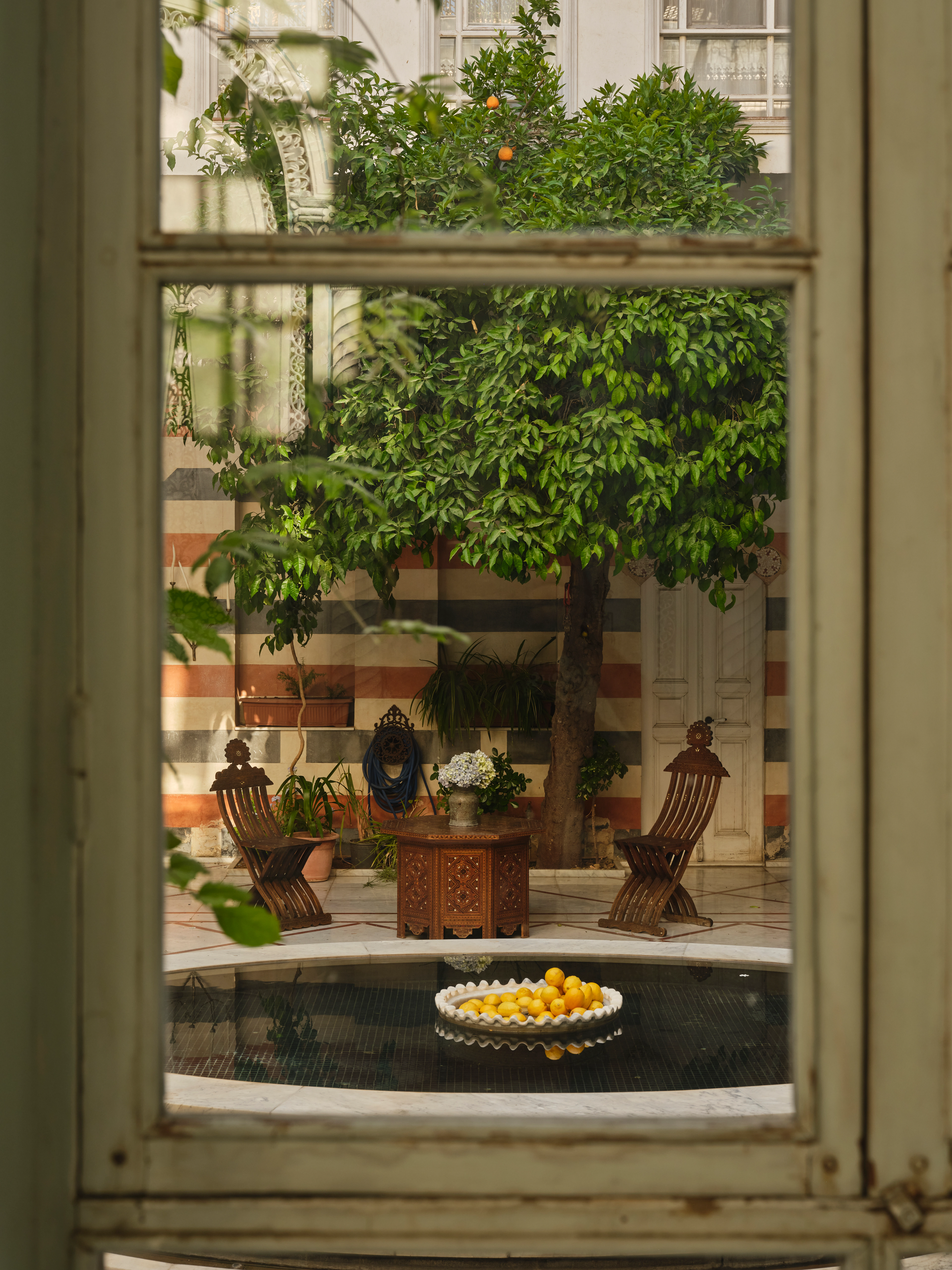 Courtyard garden viewed through a window, with citrus trees, carved wooden chairs, a small table, and a shallow pool.